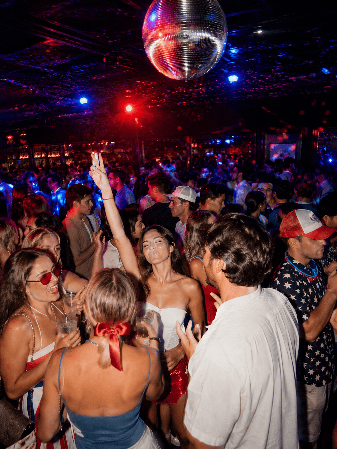 A crowded nightclub scene with people dancing and socializing under a disco ball, colorful red and blue lights illuminating the space, and a woman in the center raising her arm, smiling amidst the lively atmosphere.
