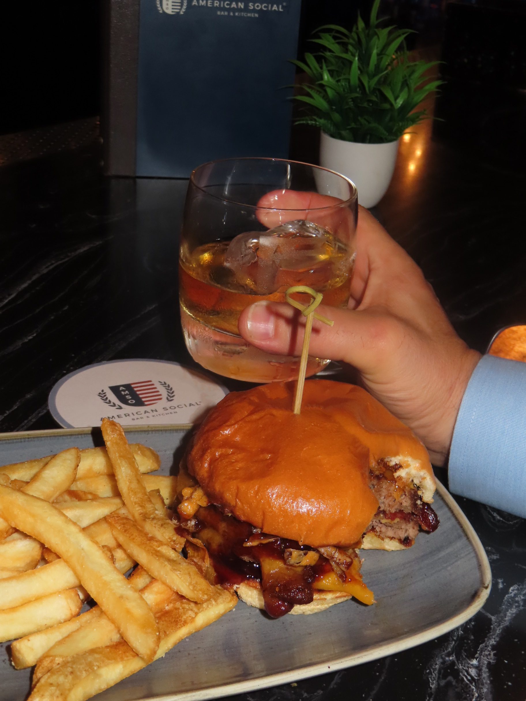 A hand holding a glass of whiskey with ice next to a burger and fries on a plate, set on a dark table with a coaster, menu, and small potted plant—perfect fuel for an evening celebrating the F1 Miami Grand Prix.