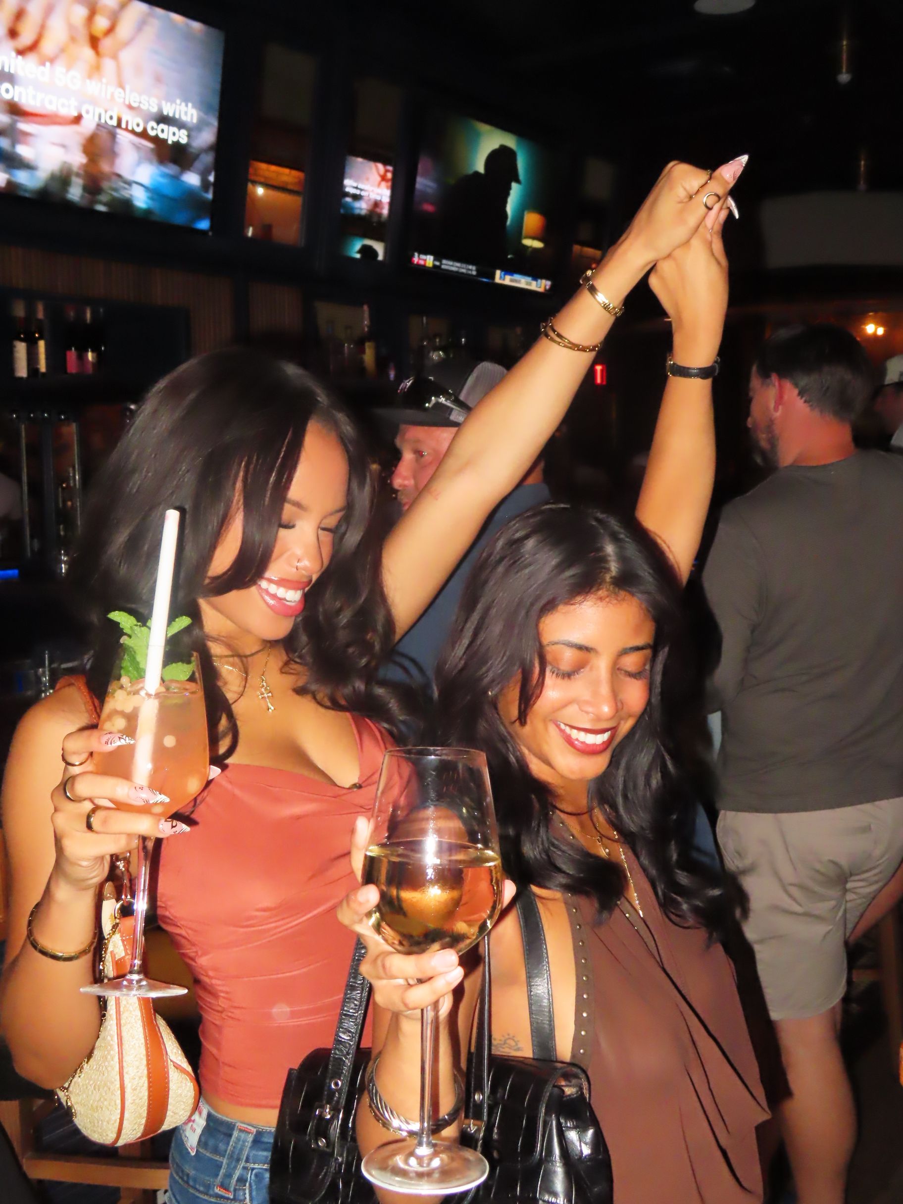 Two women smile and dance at American Social, one holding a pink cocktail with a straw and mint, the other with a glass of white wine. Lively crowds and TV screens fill the energetic bar scene in the background.