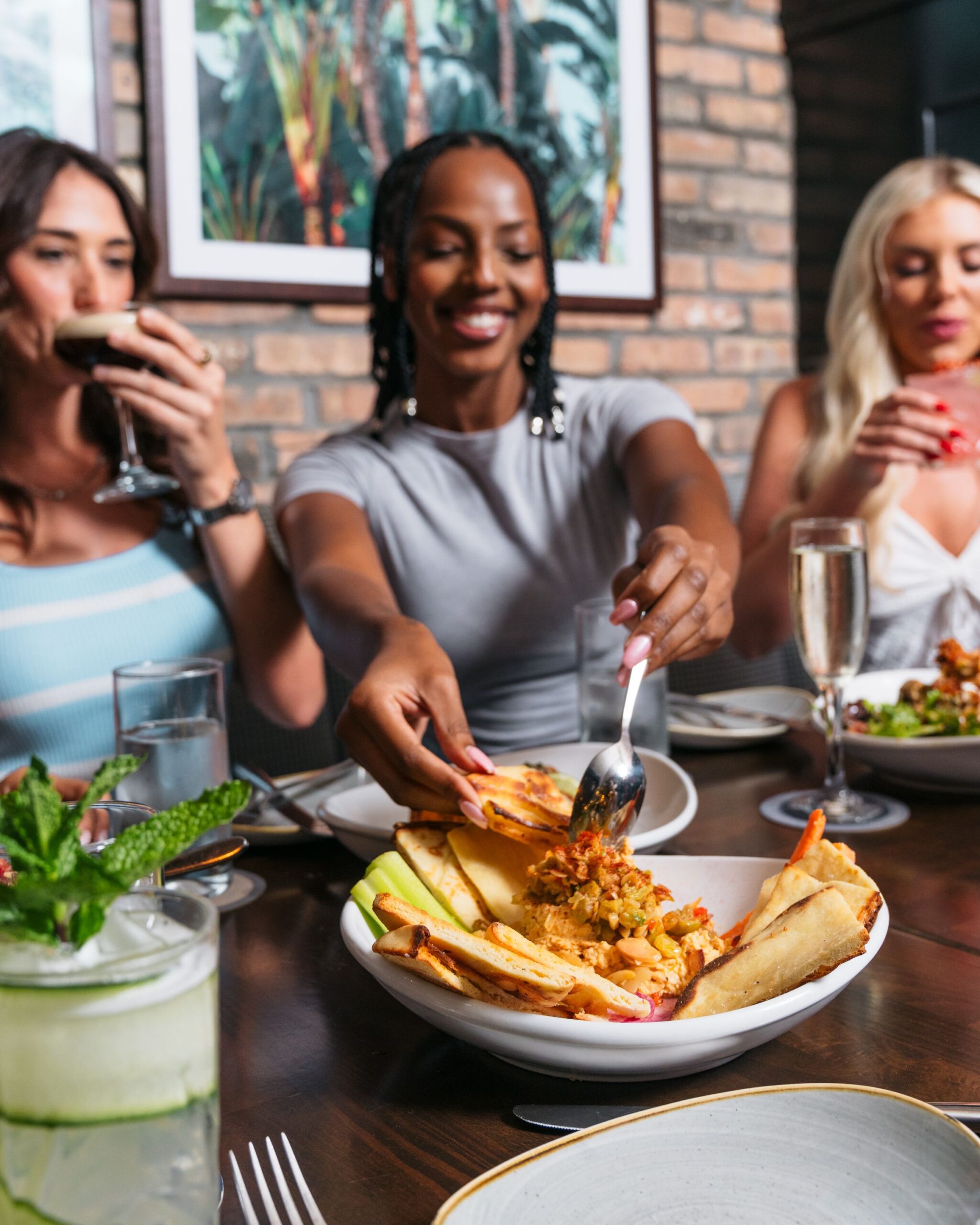 Three women sit at a restaurant table, enjoying drinks and sharing a plate of loaded fries, with one woman smiling as she serves herself. Drinks and food are visible, and a framed picture hangs on the brick wall behind them.