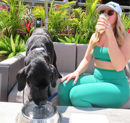 A woman in green activewear drinks from a glass while sitting on an outdoor couch. Beside her, a black dog drinks from a metal bowl. Green plants are in the background.