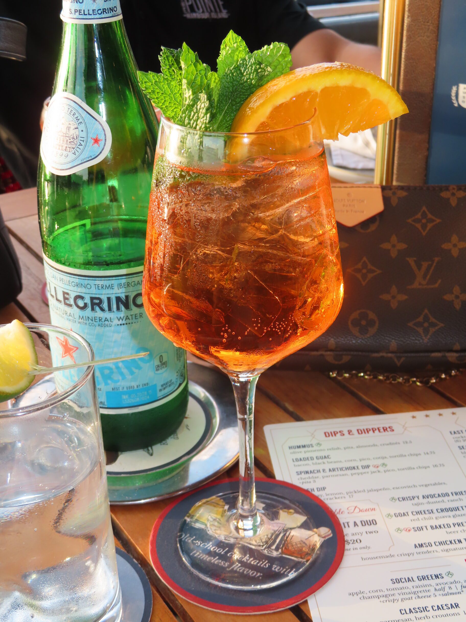 A glass of orange-colored cocktail with ice, garnished with a mint sprig and orange slice, sits on a table beside a bottle of San Pellegrino, a glass of water, menus, and a brown designer handbag.