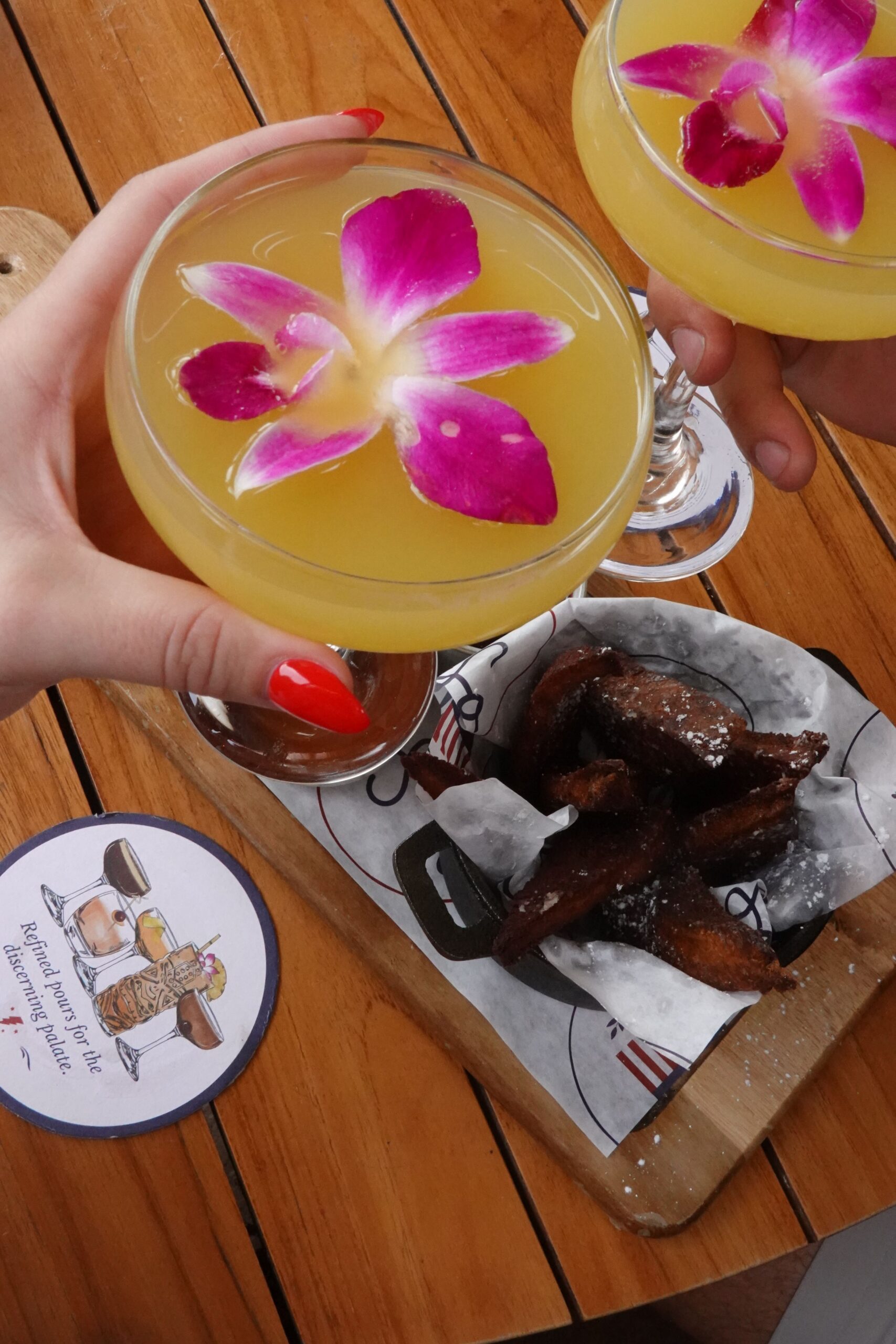 Two hands holding yellow cocktails garnished with pink orchids above a wooden table, next to a tray of fried pastries dusted with powdered sugar and a coaster with an illustration and text.