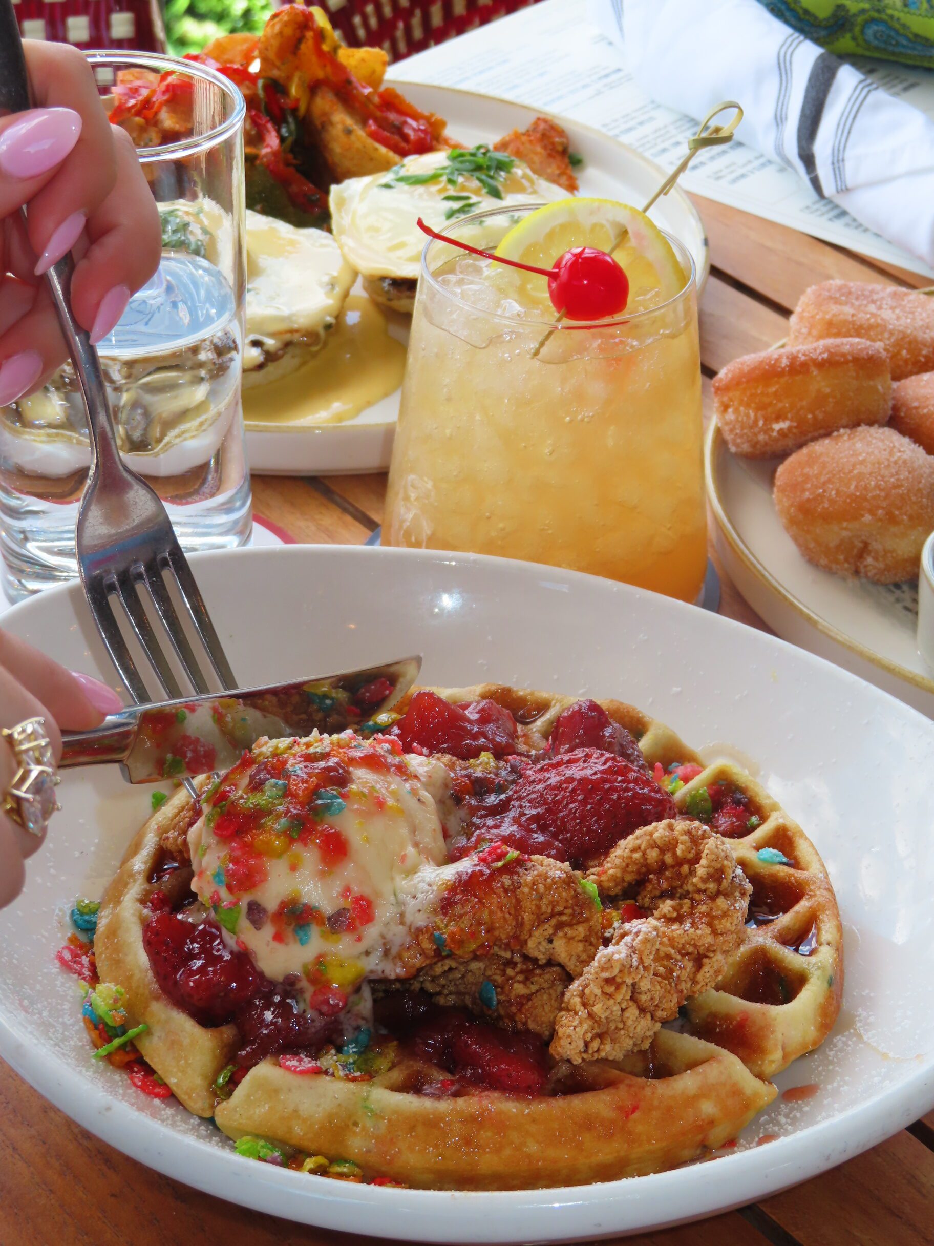 A hand cuts into a waffle topped with fried chicken, ice cream, fruit sauce, and colorful cereal. On the table are a drink with a cherry and lemon, donuts, water, and more brunch dishes in the background.