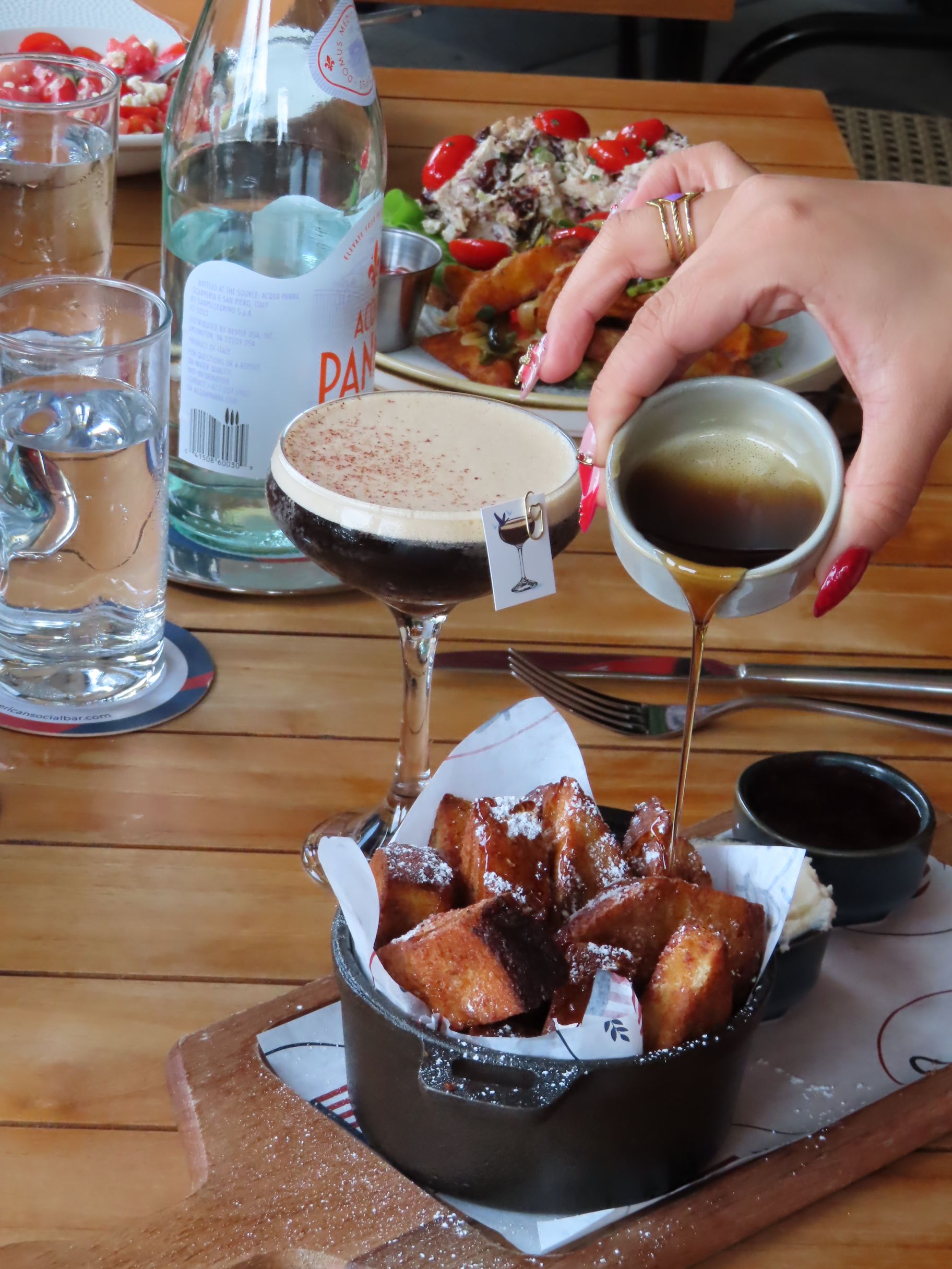 A hand pours sauce over a bowl of fried pastries on a wooden table, next to a glass of espresso martini, water glasses, and plates of food in the background.