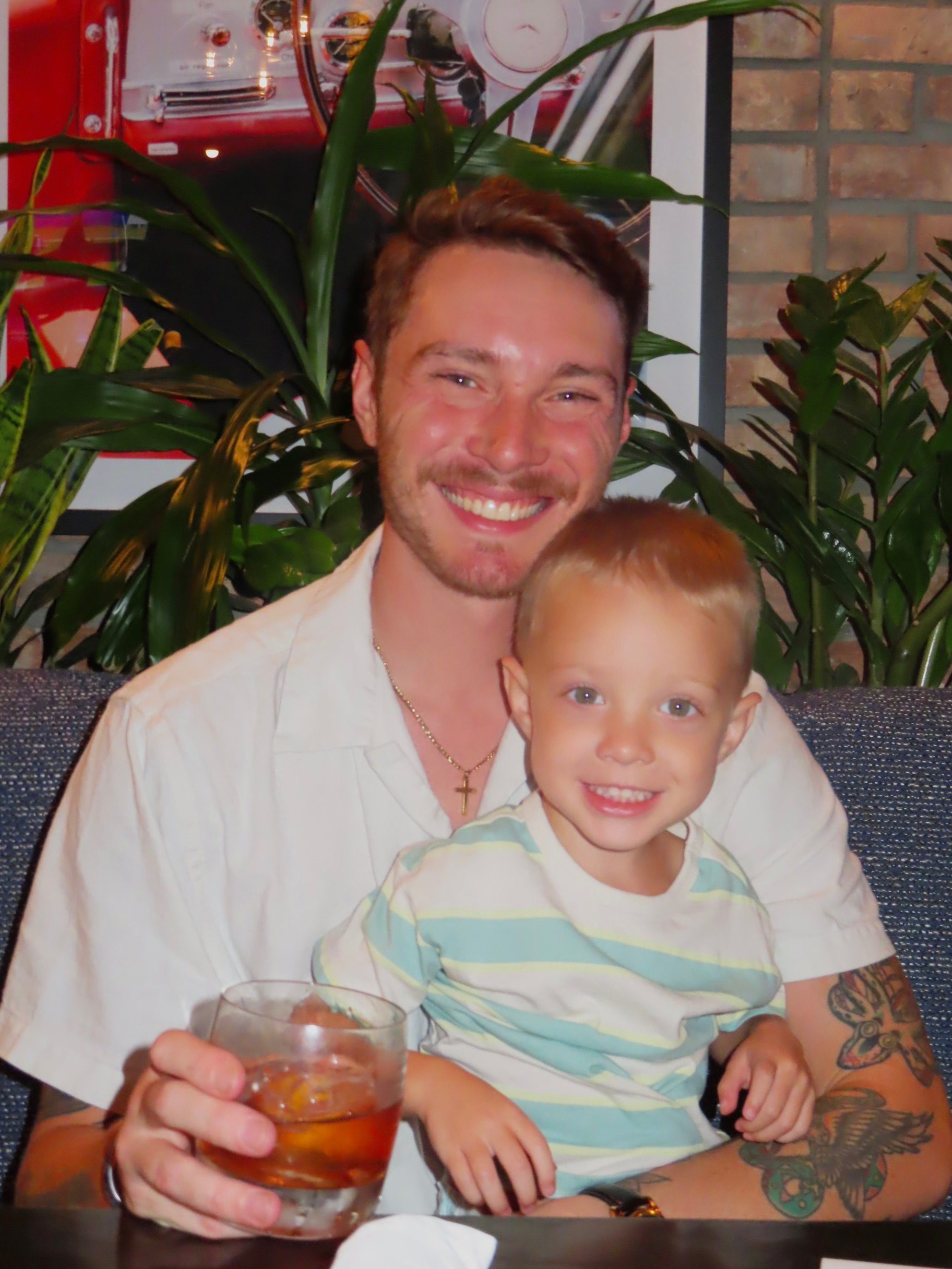 A smiling man with a drink in his hand sits next to a young boy who is also smiling. They are seated indoors on a bench with green plants and a brick wall in the background.