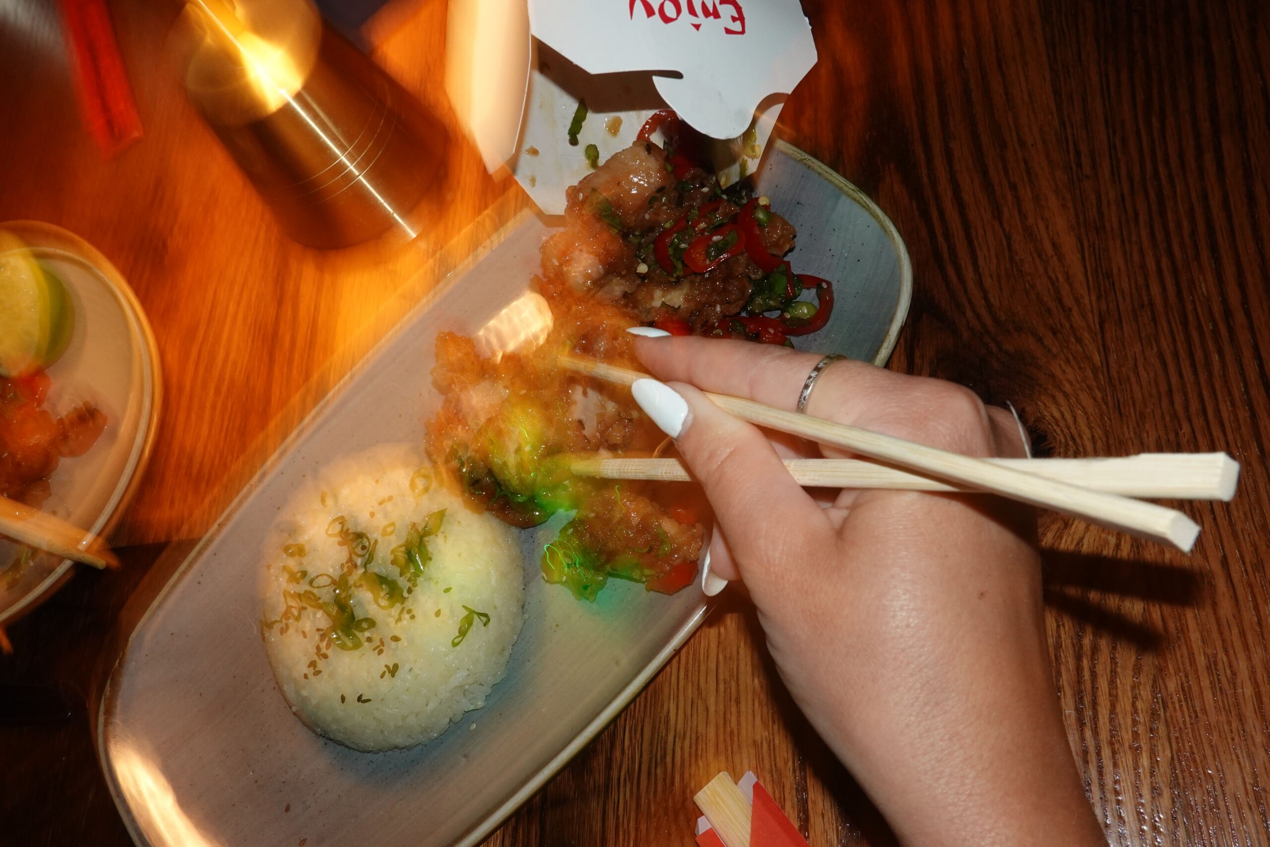 A hand holding chopsticks prepares to pick up food from a plate with rice and stir-fry at American Social. A takeout container and other dining items are on a wooden table under warm lighting.