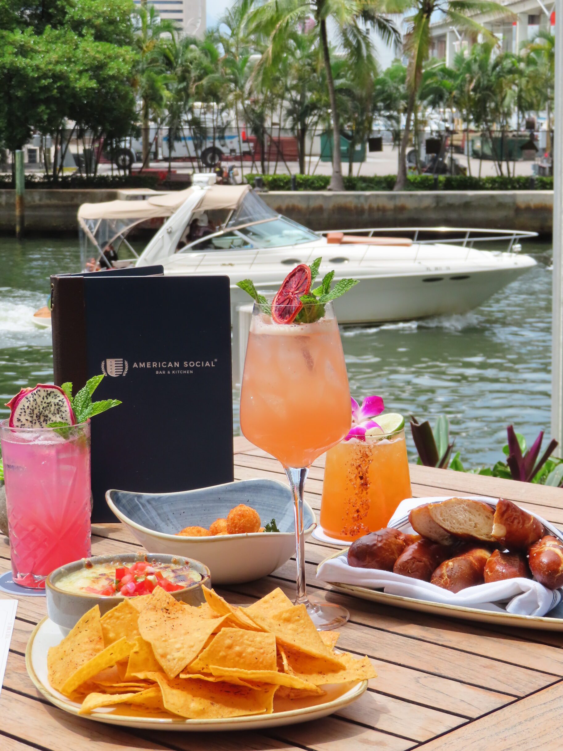 A table with cocktails, nachos, pretzels, and appetizers is set outdoors by the water. A boat passes by in the background, and palm trees line the waterfront. A restaurant menu is on the table.