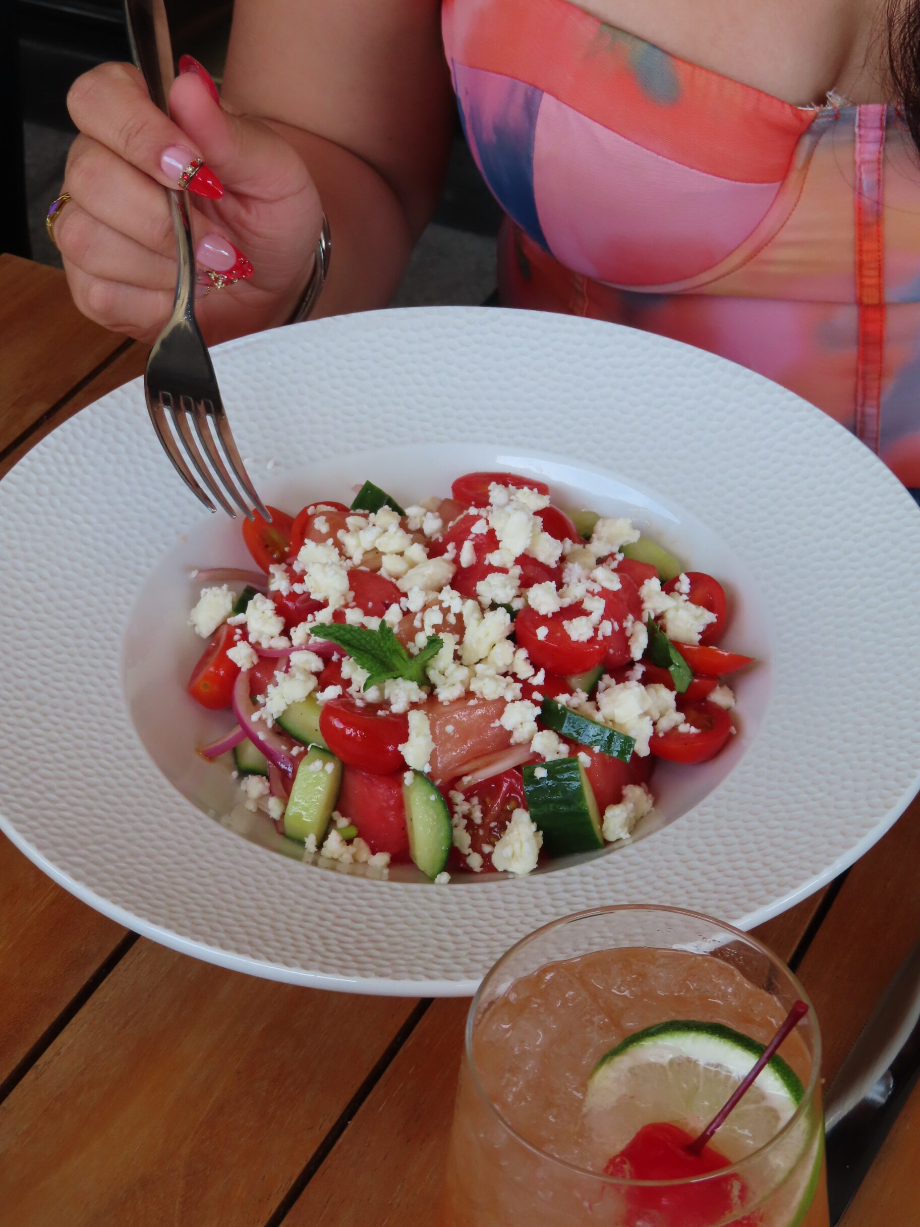 A person wearing a colorful strapless dress holds a fork above a bowl of Greek salad with tomatoes, cucumbers, onions, and feta cheese. A drink with ice, lime, and a cherry is on the wooden table.
