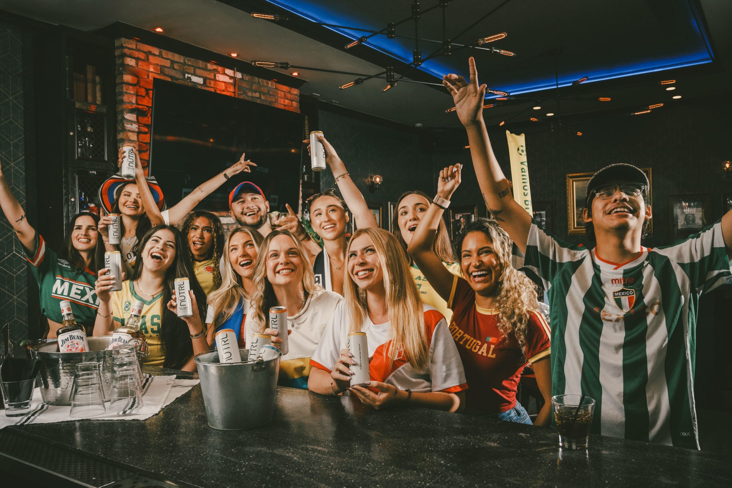 A group of excited young adults in sports jerseys cheer and hold drinks at a bar, celebrating together. Some have their arms raised, and everyone is smiling. There are bottles and an ice bucket on the counter.