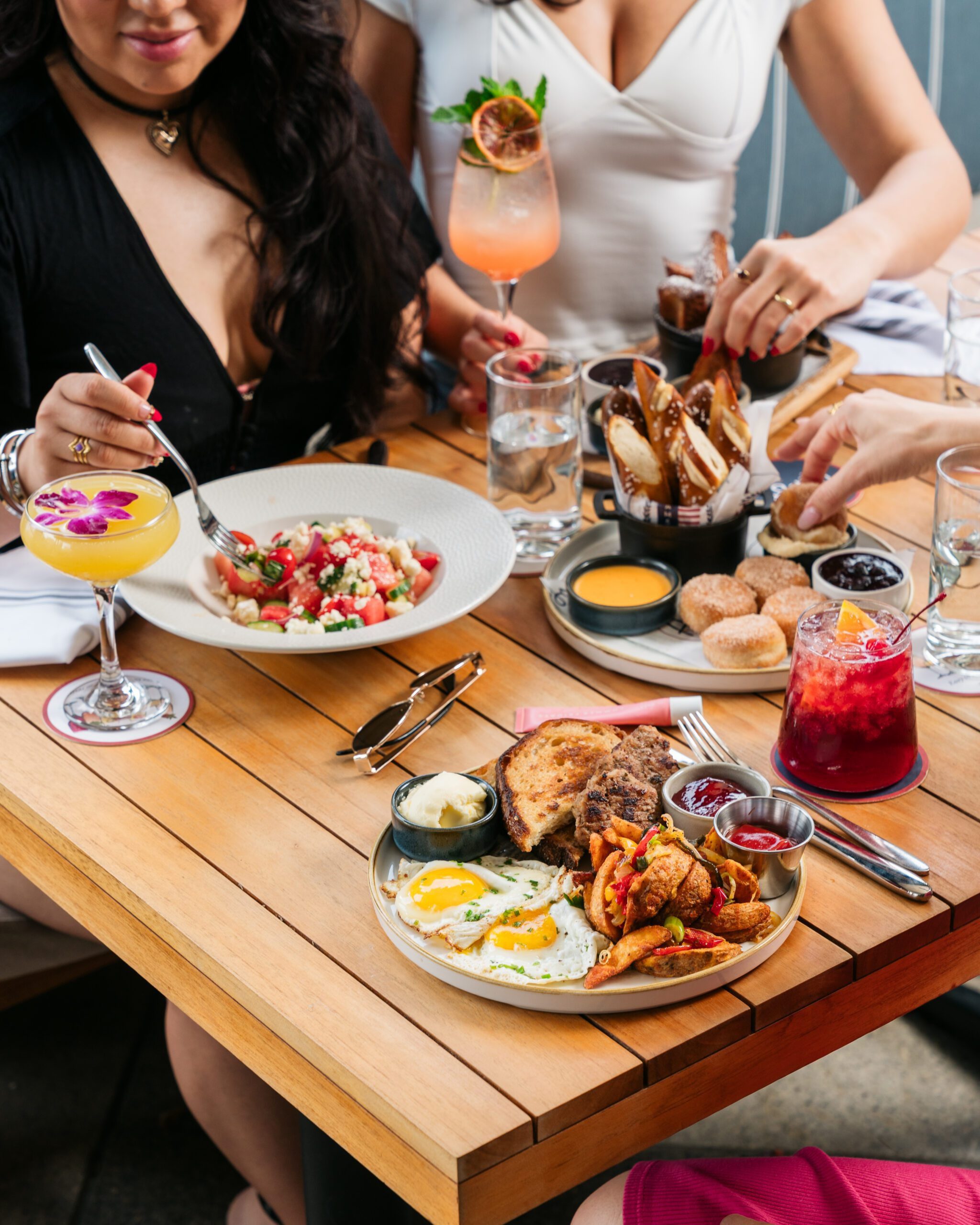 Three women sit at a wooden table enjoying brunch dishes and colorful drinks, including fried chicken with eggs, a salad, toast with spreads, and cocktails. Hands reach for food, creating a lively, social scene.