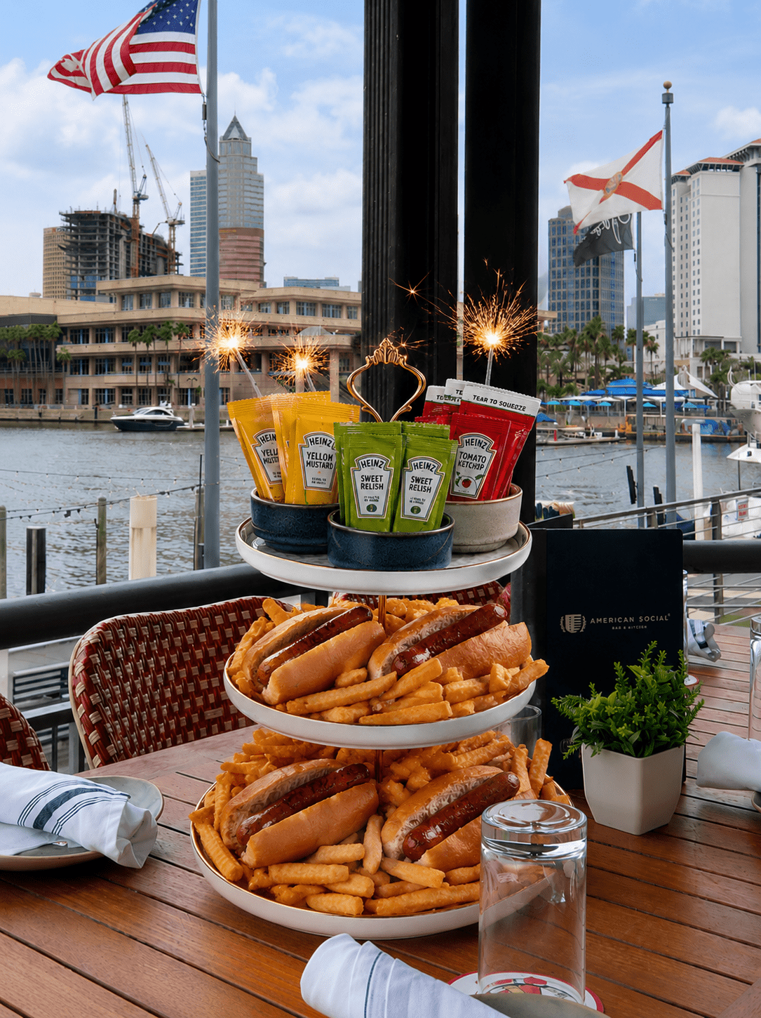 A tiered tray holding hot dogs and fries sits on an outdoor table by the water, with city buildings and American flags in the background. Sparklers and assorted ketchup and mustard bottles top the tray.