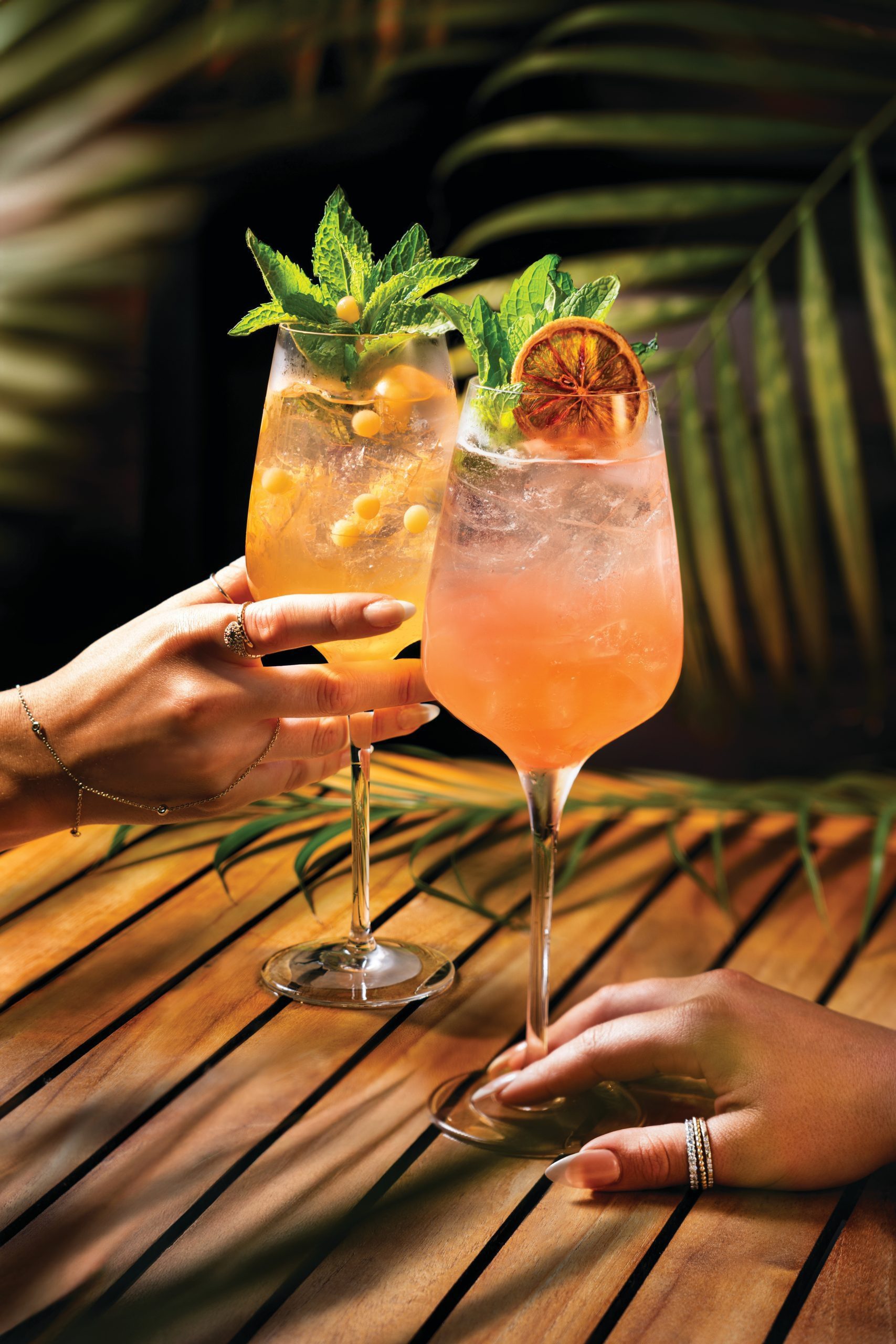Two hands holding elegant cocktail glasses filled with colorful, refreshing drinks garnished with mint leaves, berries, and a dried citrus slice, set on a wooden table with tropical leaves in the background.