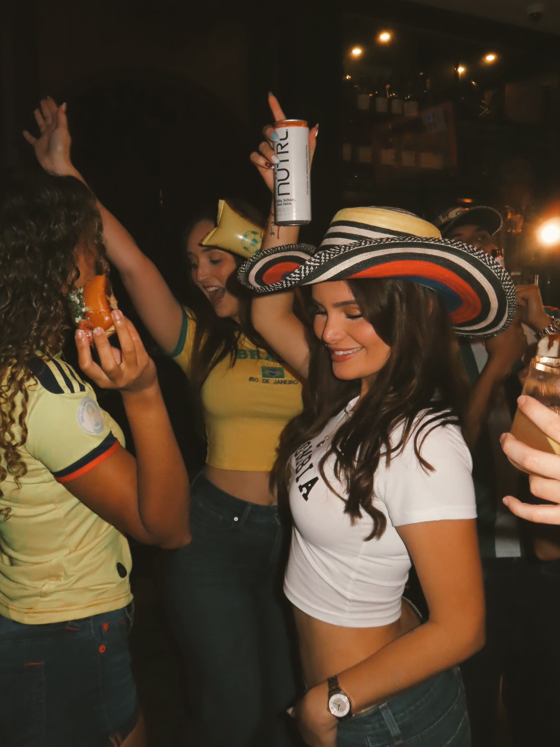 Three young women dance and celebrate at a lively party during the F1 Miami Grand Prix. One wears a wide-brimmed hat and holds a drink can, while another raises her arms and smiles amid the festive atmosphere.