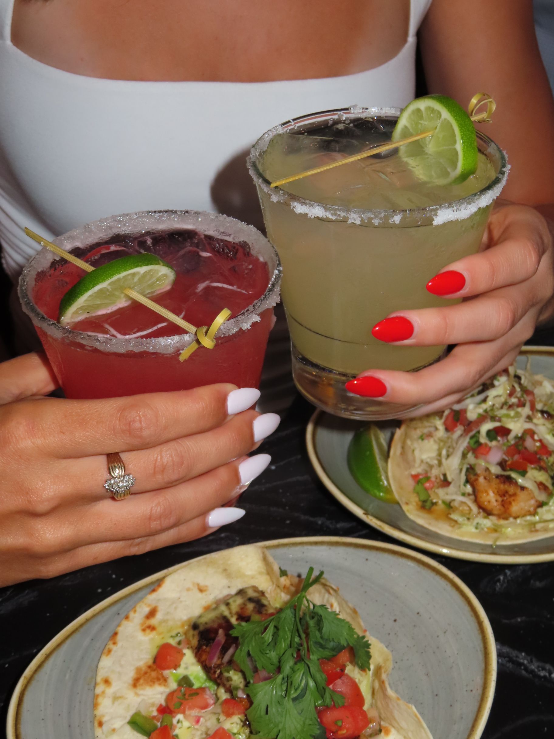 A woman with manicured nails holds two cocktails—one red, one yellow—garnished with lime wedges. Plates of tacos topped with herbs, vegetables, and fish are on the table in front of her.