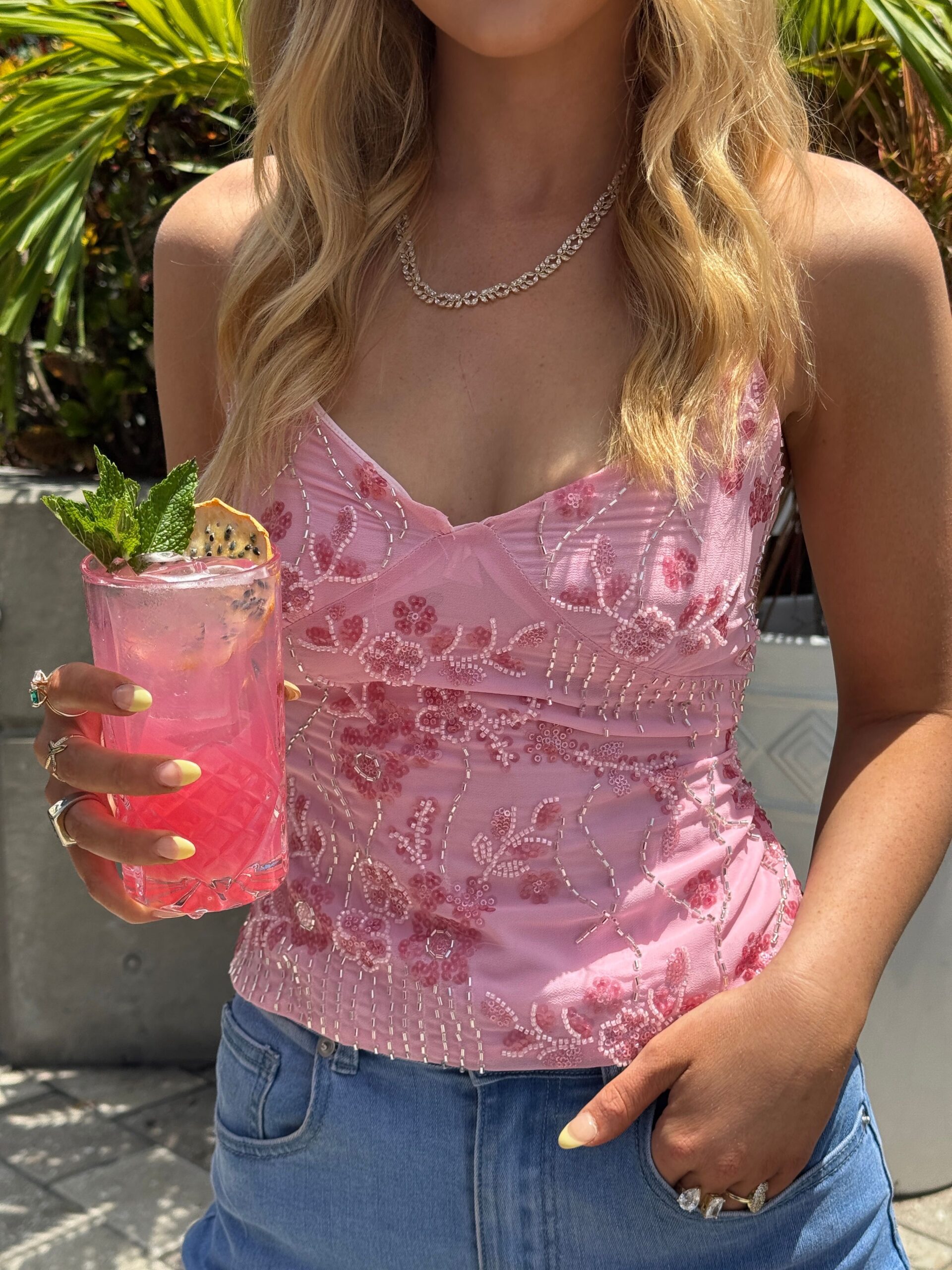 A woman in a pink, floral, beaded top and blue jeans holds a bright pink cocktail with mint and a dragon fruit slice garnish. She stands outdoors in sunlight near green plants.