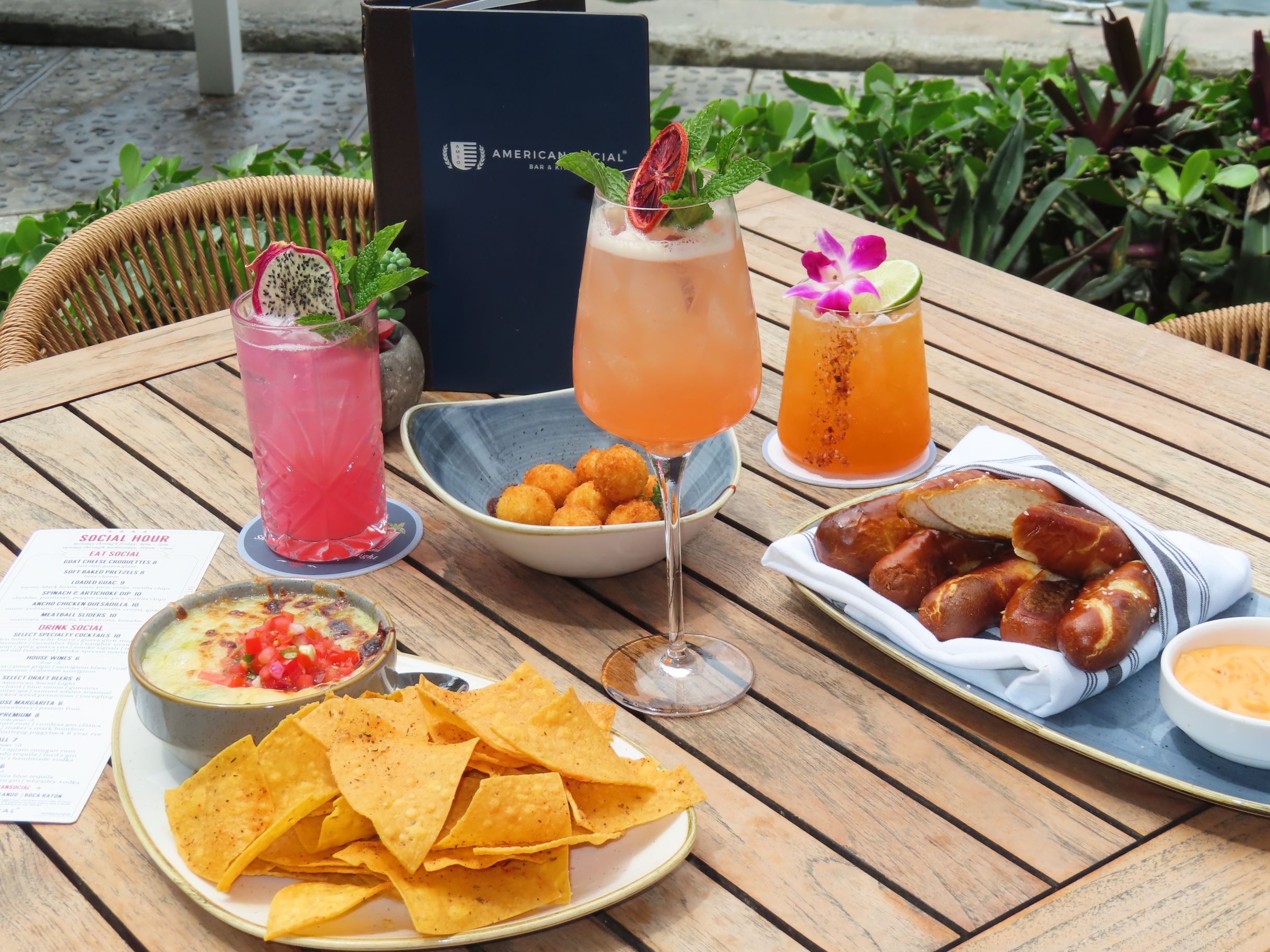 A wooden table at American Social set with three colorful cocktails, a plate of nachos with dip, a bowl of pretzel bites, a dish of fried cheese balls, and a menu, with lush greenery in the background.