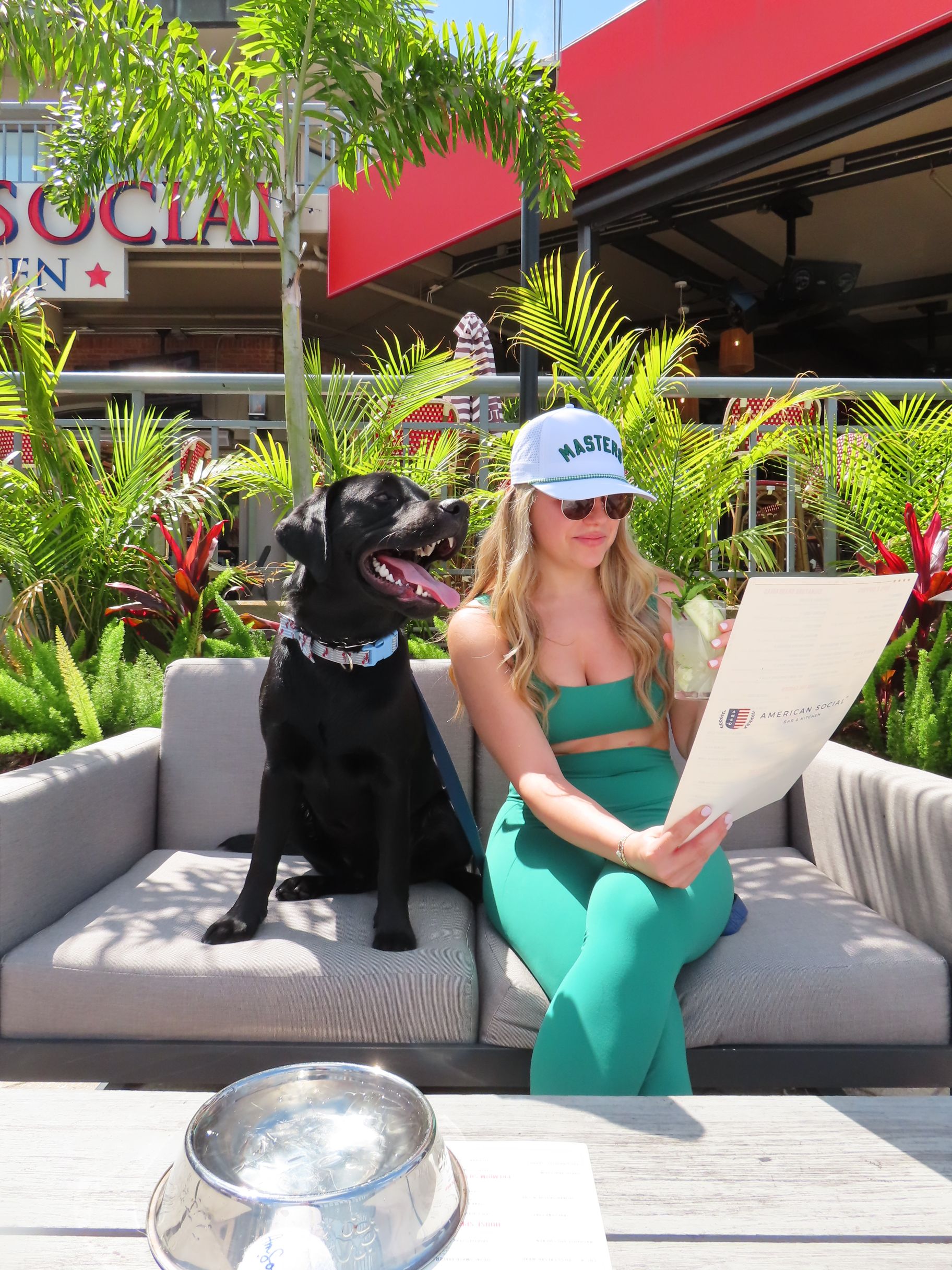 A woman in a green outfit and white cap sits on an outdoor couch with her black dog beside her at American Social. She reads a menu while the dog, looking happy with its mouth open, enjoys the lush plants and vibrant restaurant scene around them.