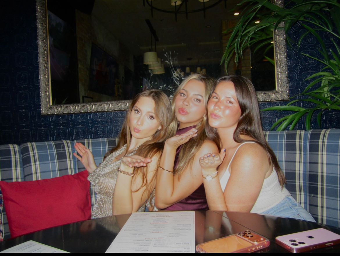 Three young women sit on a plaid couch at American Social, posing for the camera with puckered lips and blowing kisses. Surrounded by menus and a plant, they enjoy the lively vibe with a large mirror reflecting the restaurant behind them.
