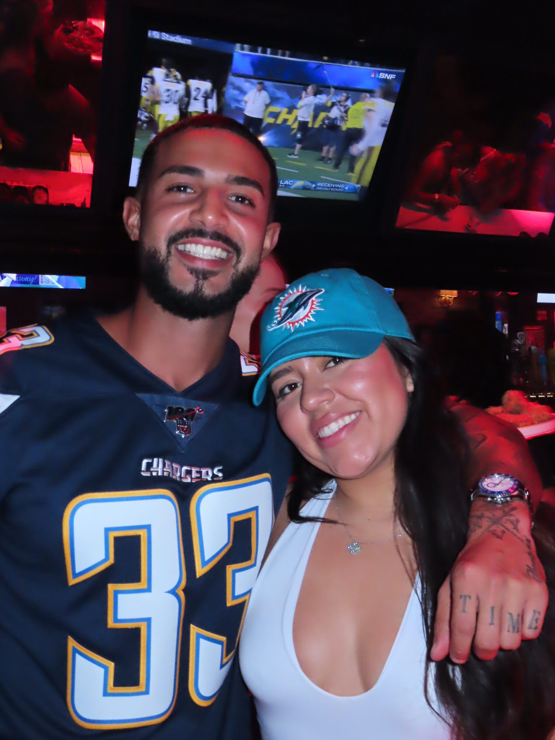 A smiling man in a Chargers jersey hugs a woman in a Dolphins cap. A football game plays on a TV screen in the background.