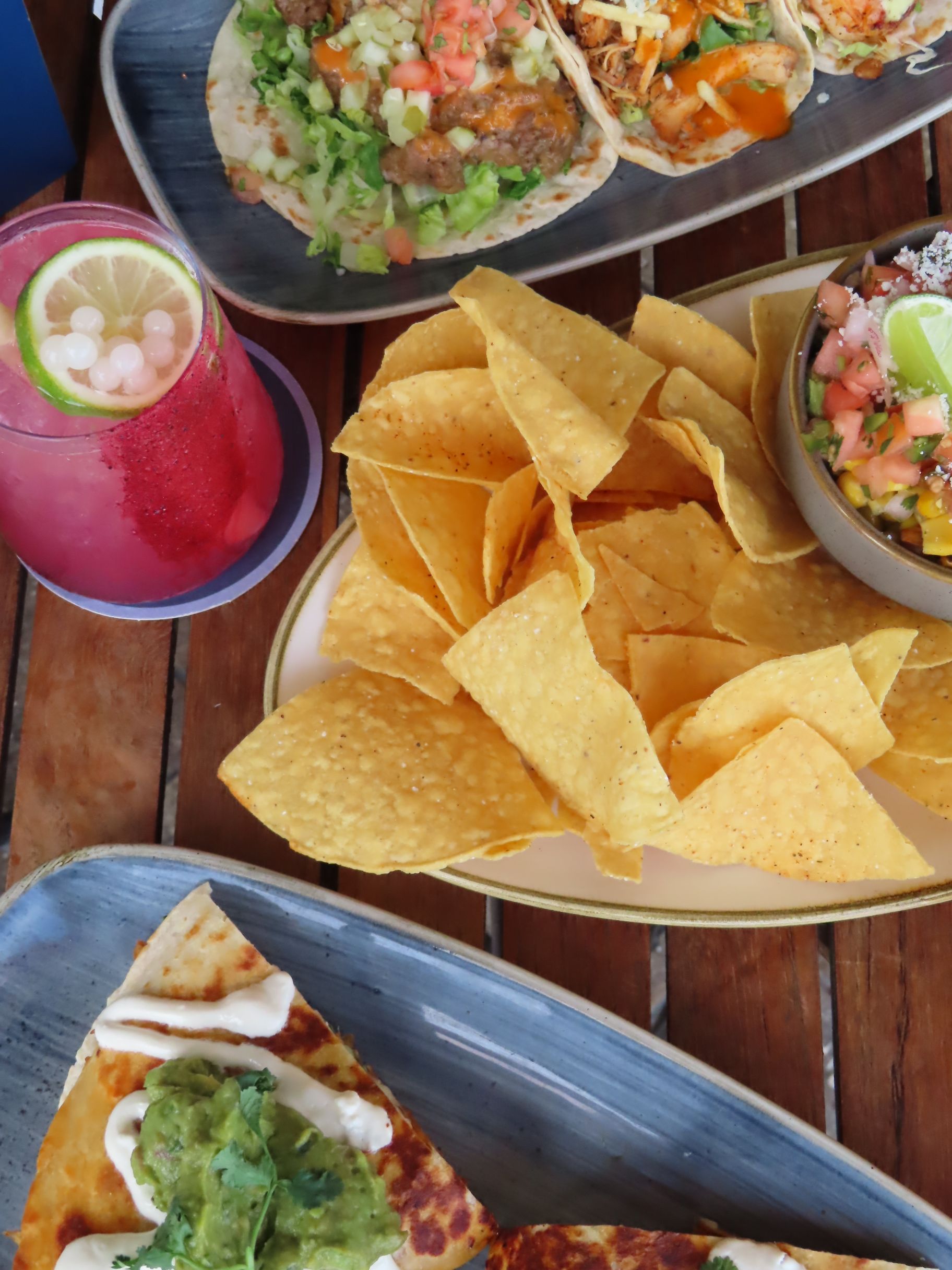 A plate of tortilla chips, tacos, a pink drink with ice and lime, salsa with lime, and a slice of pizza with guacamole and sour cream, all on a wooden table.