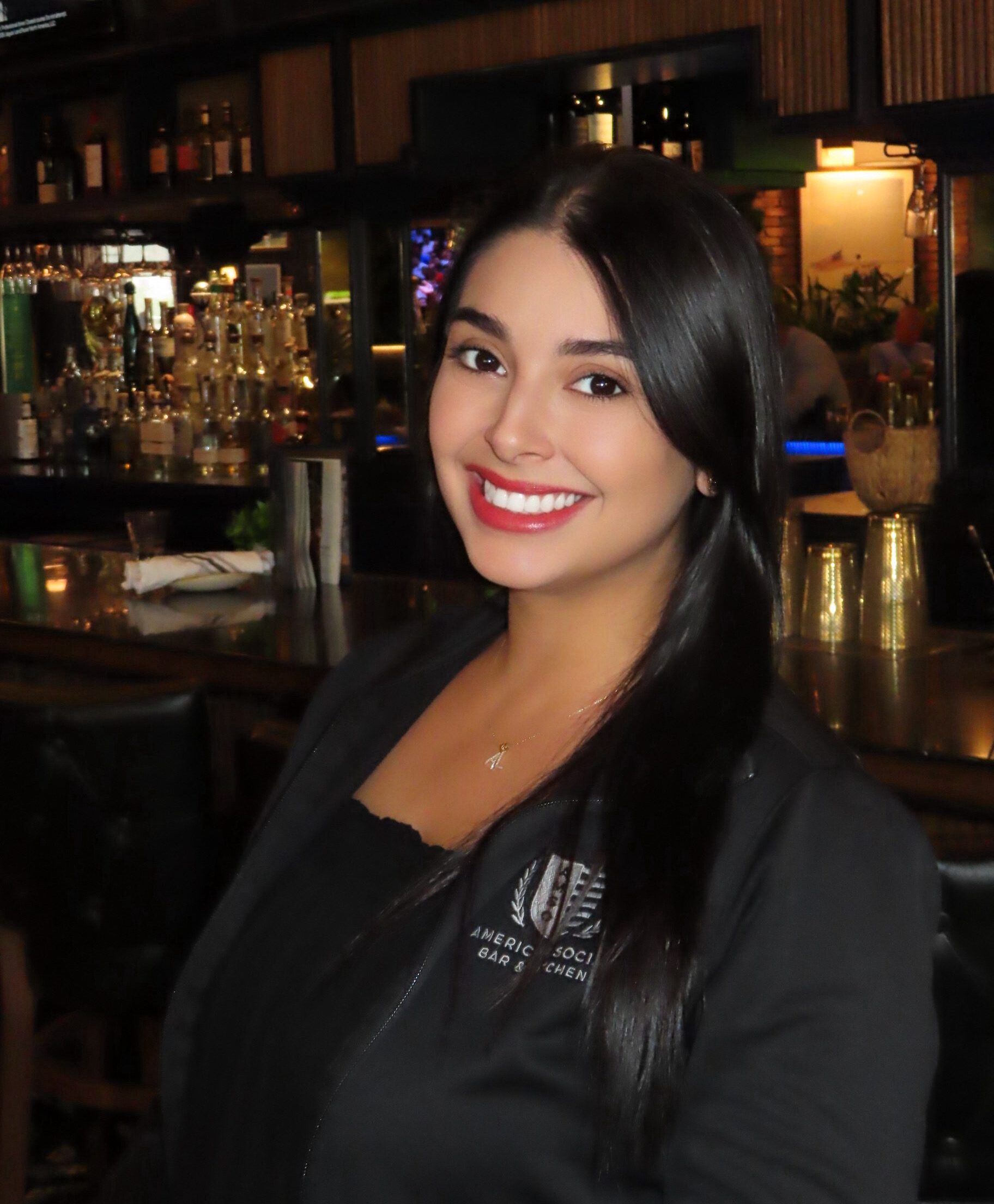 A woman with long dark hair and a bright smile sits at a bar with shelves of glasses and bottles behind her. She wears a black top and dark jacket. Televisions are mounted above the bar.
