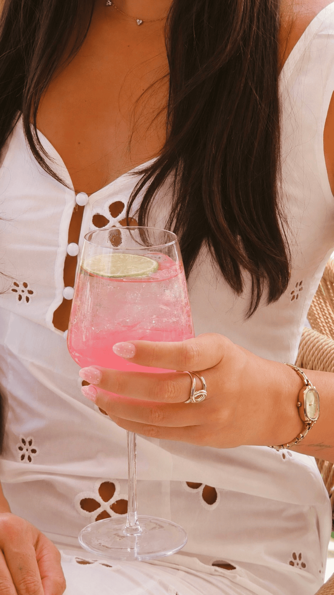 A woman in a white eyelet dress holds a glass of pink drink with ice and a lime slice. She wears a gold watch, bracelet, ring, and a delicate necklace.