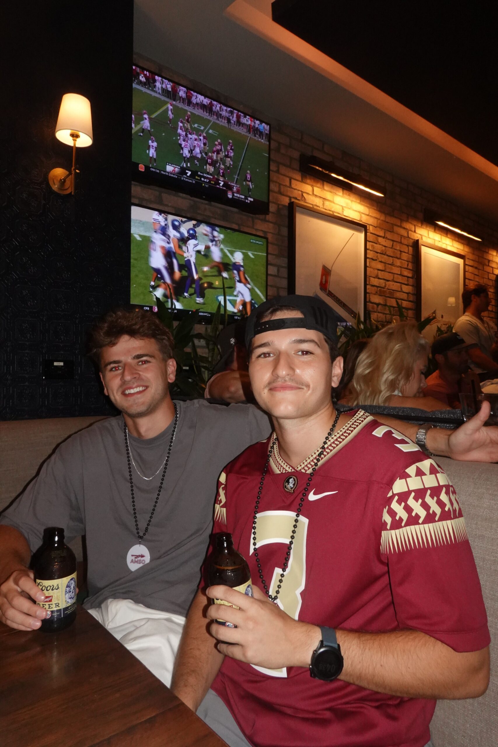 Two young men sit at a table in a bar, both holding bottled drinks. One wears a Florida State jersey. Football games are playing on TVs in the background, and other people are seated nearby.