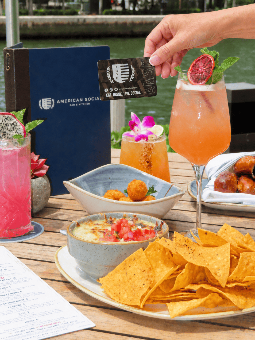 A table with cocktails, nachos with dip, appetizers, and a menu at an outdoor restaurant. A hand holds a loyalty card near a cocktail garnished with a dried orange slice and mint.