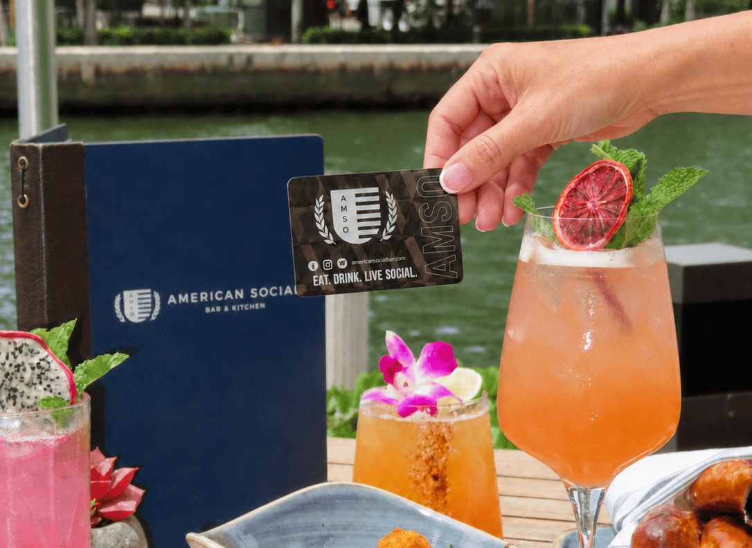 A hand holds a card above a cocktail surrounded by colorful drinks, chips with dip, fried appetizers, and an American Social menu on a wooden outdoor table by the water.