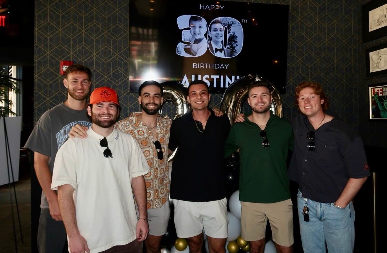 Six smiling men stand together indoors in front of a “Happy 30th Birthday Austin” sign with balloons. They are dressed casually, and the setting appears to be a lively birthday event celebrating a special milestone.