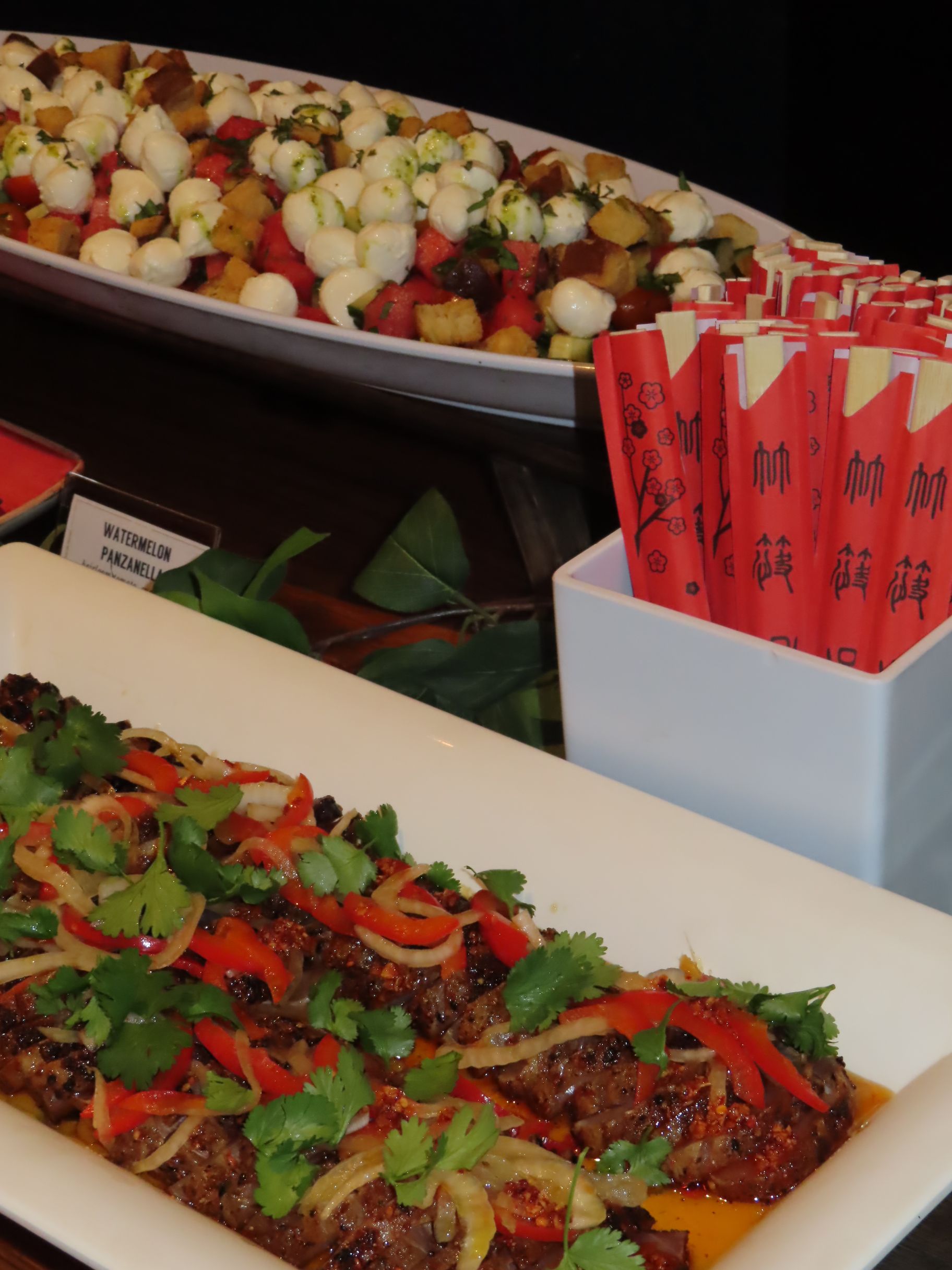 A platter of grilled meat topped with herbs and peppers, a bowl of watermelon salad with mozzarella balls, and a container of red chopsticks are displayed on a table—perfect for serving at events or special gatherings.