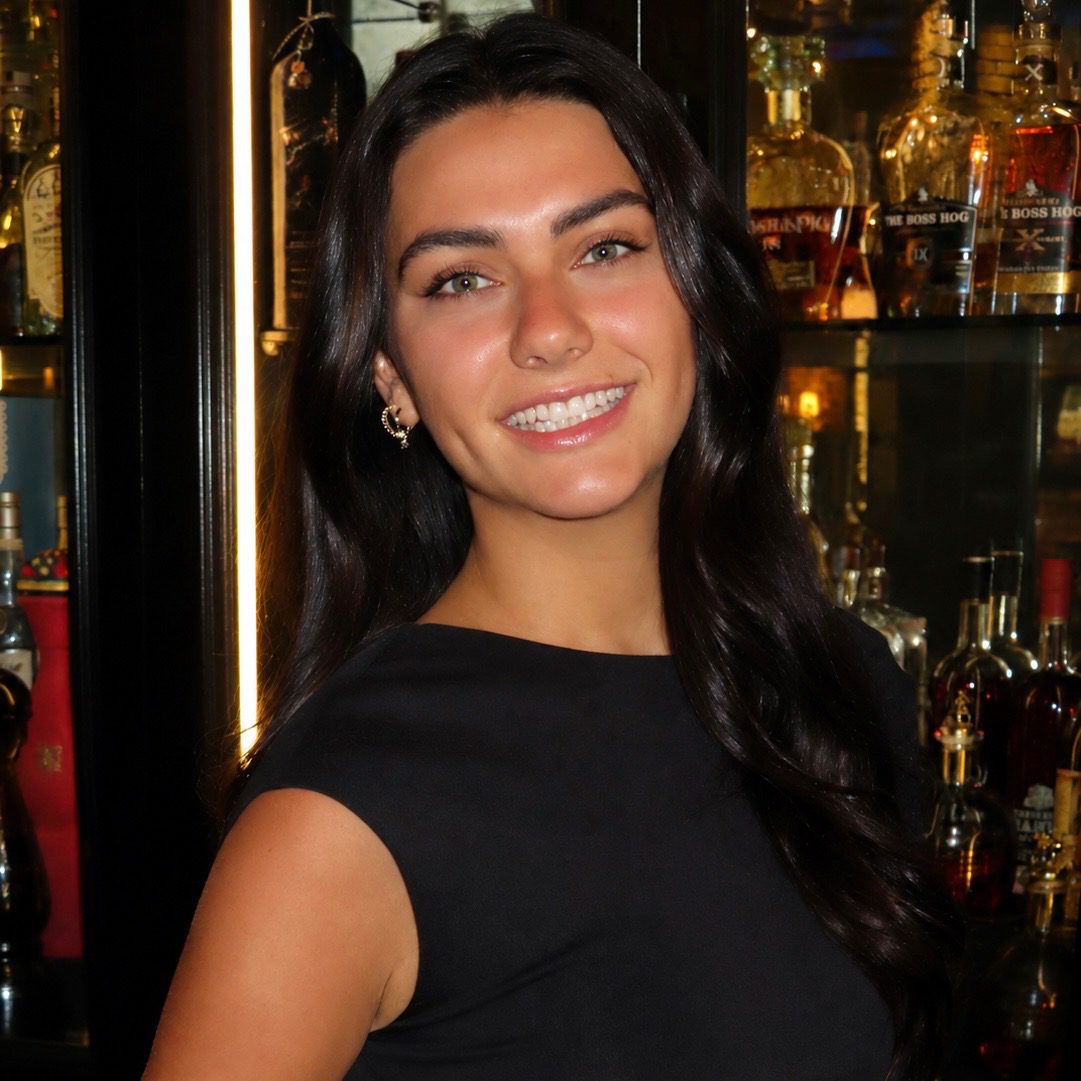 A woman with long dark hair and a black sleeveless dress smiles in front of a display case filled with various bottles of alcohol.
