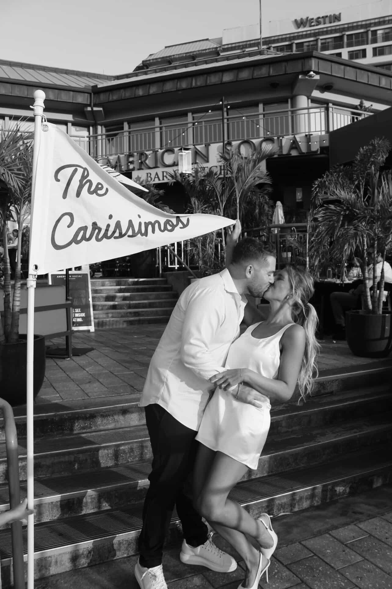 A couple in white outfits kisses on outdoor steps during special events, holding a flag that reads “The Carissimos.” Palm plants and a building with “Westin” and “American Social” signs are visible in the background.
