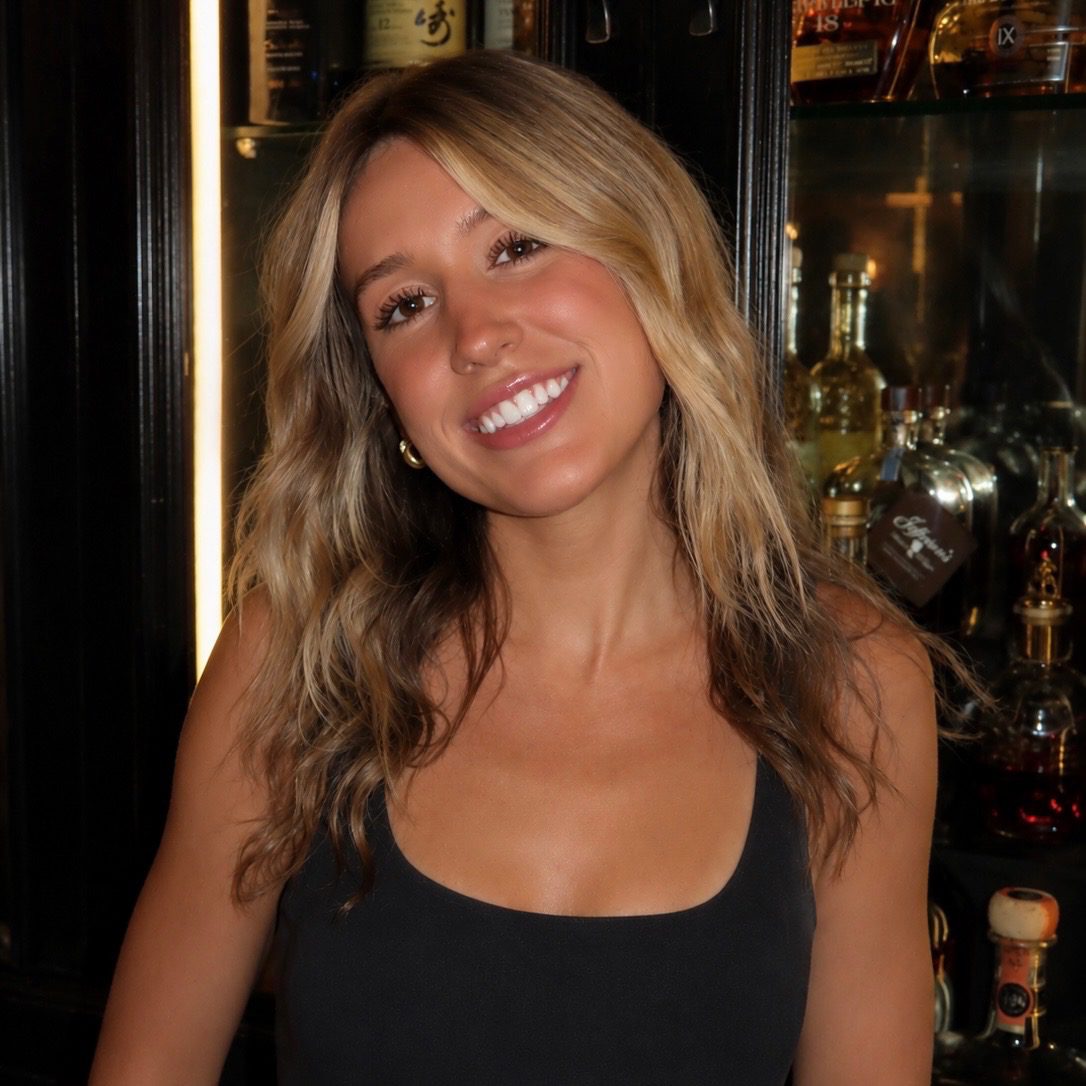 A woman with wavy blonde hair and a black top smiles in front of a bar shelf filled with bottles.