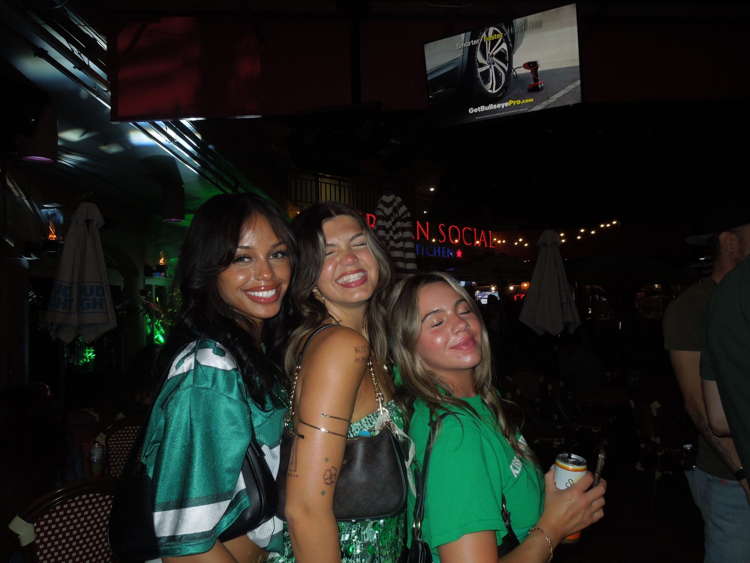 Three women smiling and posing together at a lively nighttime event. They are wearing green outfits and look happy. Bright lights and outdoor seating are visible in the background, capturing the vibrant atmosphere of such memorable events.