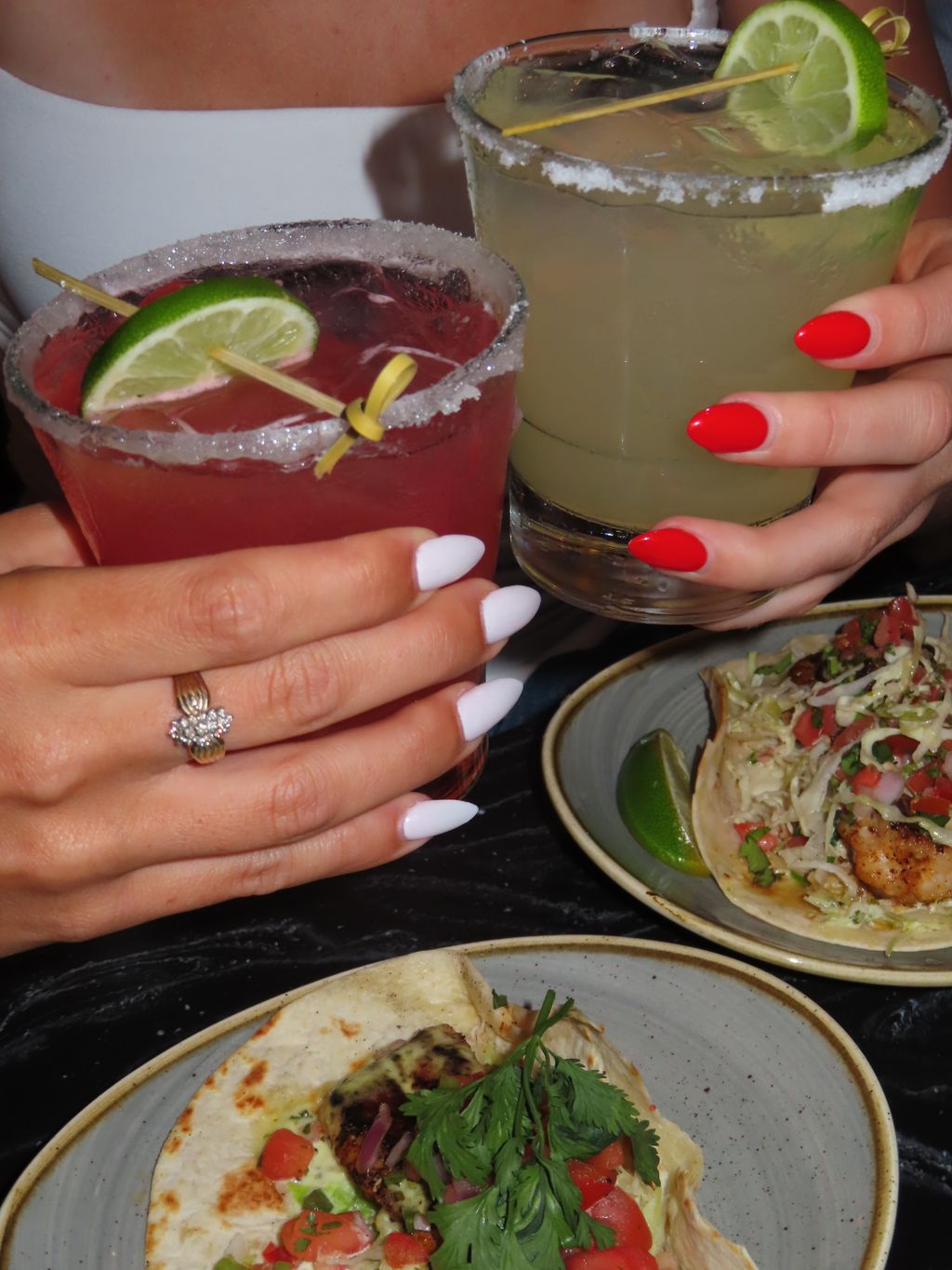 Two hands with manicured nails hold margarita glasses, one pink and one yellow, above plates of tacos garnished with greens, tomatoes, and lime wedges on a dark table.