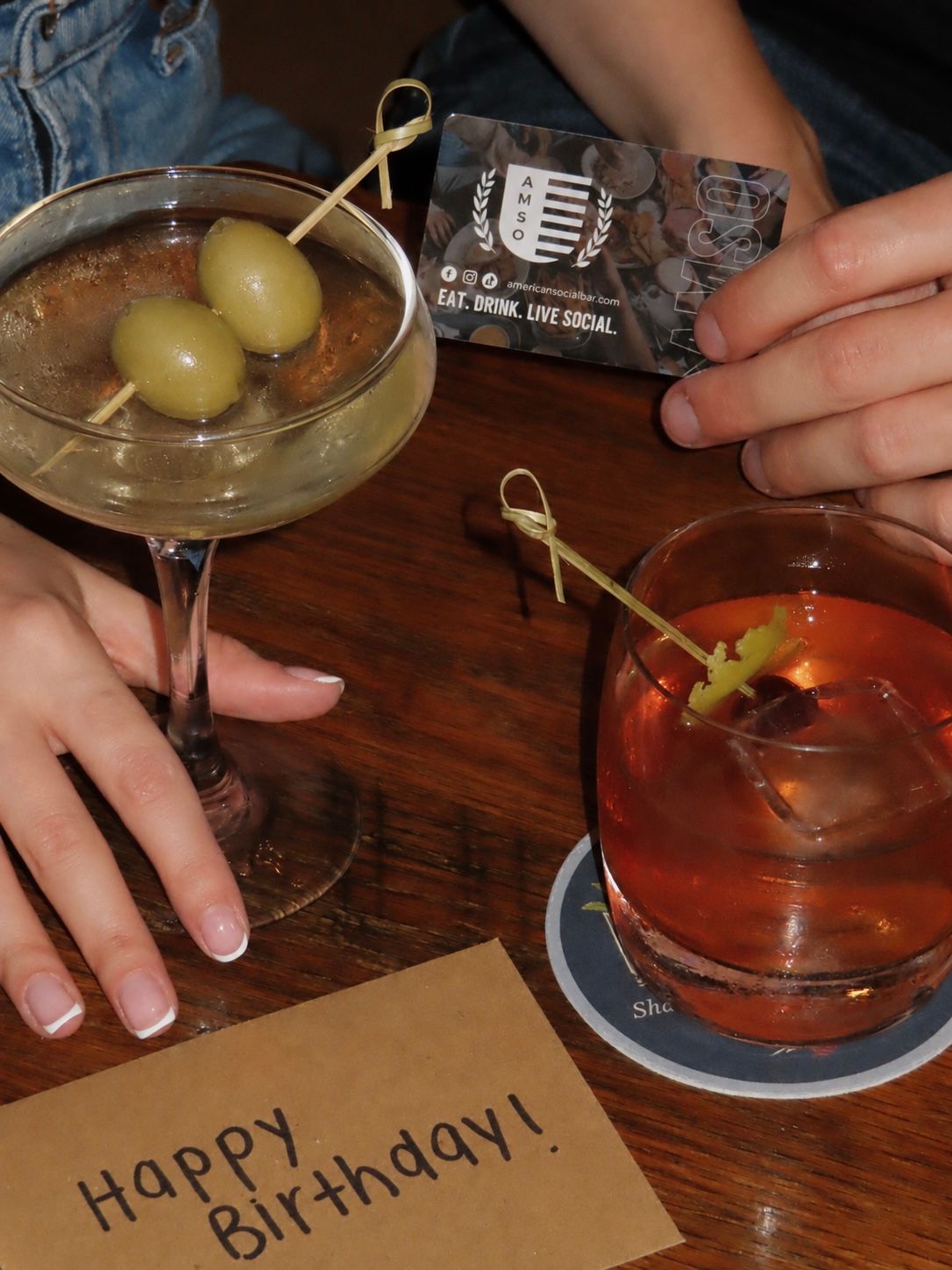 Two hands holding cocktails—one martini with olives and one orange drink—on a wooden table. A card, a coaster, and an envelope reading “Happy Birthday!” are also visible.