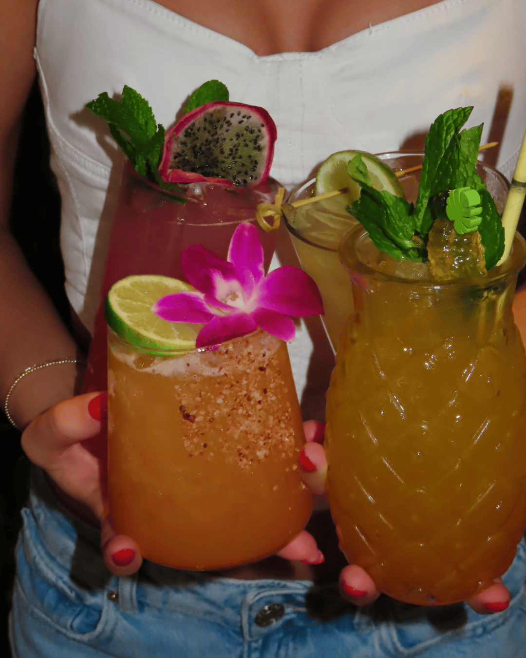 A person in a white top and jeans holds three tropical cocktails garnished with mint, lime, flowers, and fruit.