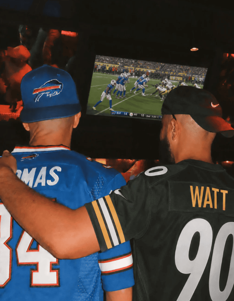 Two men wearing NFL jerseys, one for the Buffalo Bills and one for the Pittsburgh Steelers, watch a football game on a TV screen in a dimly lit room, sharing an arm draped around each other’s shoulders as they enjoy one of the season’s most exciting events.
