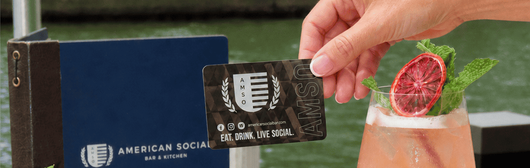 A hand holds an “American Social” card above a cocktail garnished with mint and dried citrus, with a blue menu and water in the background.