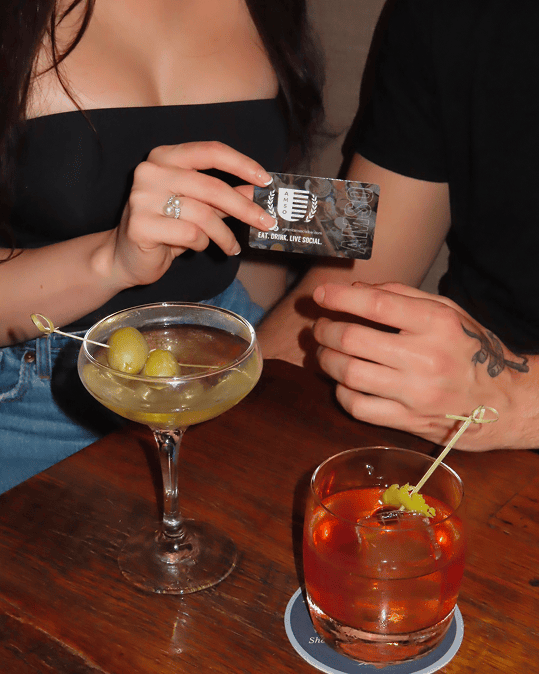 Two people sit together at a wooden table with cocktails, one holding a card that reads “EAT. DRINK. LIVE. SOCIAL.” A martini with olives and a reddish drink with a garnish are on the table.