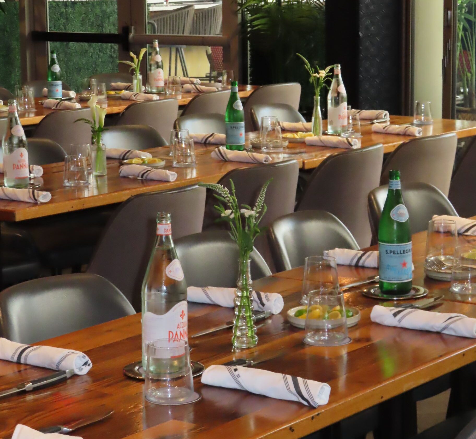 A restaurant table set with napkins, glasses, bottled water, and small vases with greenery offers an inviting atmosphere for events. Several neatly arranged chairs surround the table, making the setting appear ready to welcome guests.