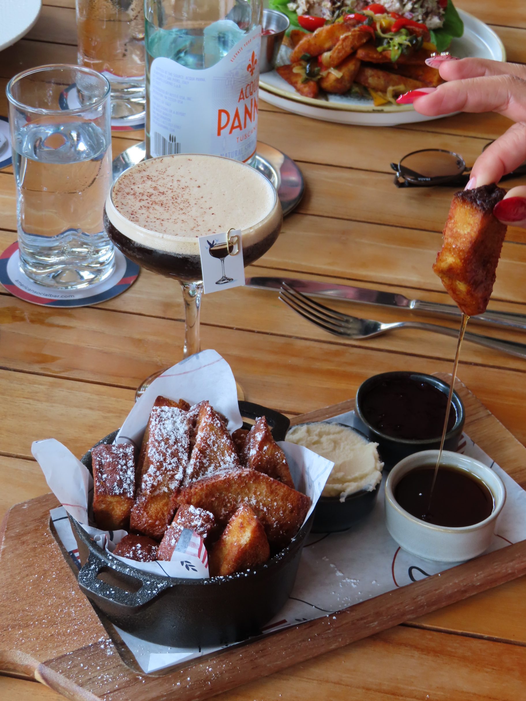 A hand dips a piece of powdered French toast into syrup. A small pot of French toast sticks, whipped butter, and syrup sit on a wooden board. An espresso martini and water are in the background.