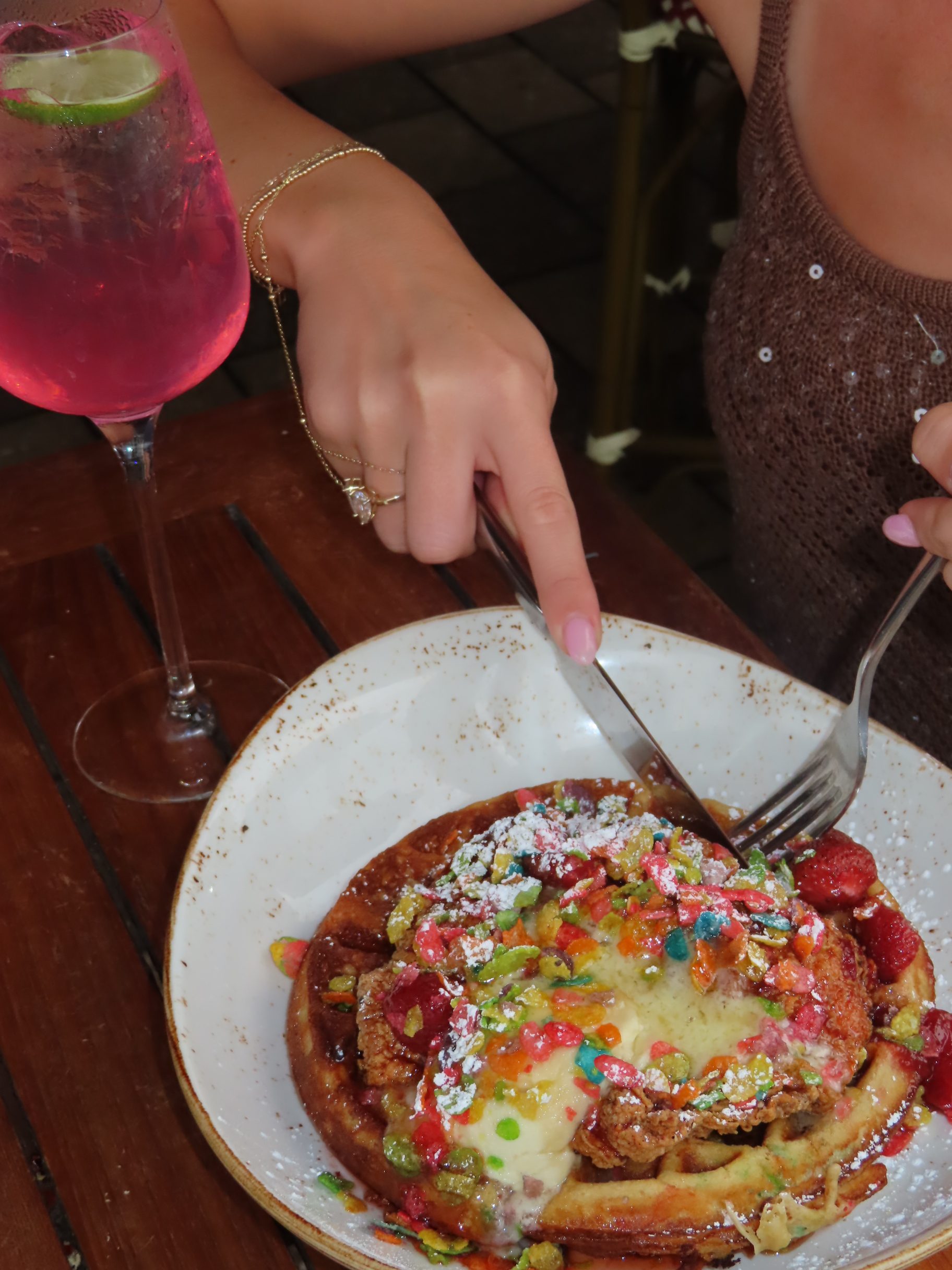 A person in a sparkly top cuts into a waffle topped with colorful cereal, strawberries, and cream. A pink sparkling drink in a wine glass sits on the wooden table beside the plate.