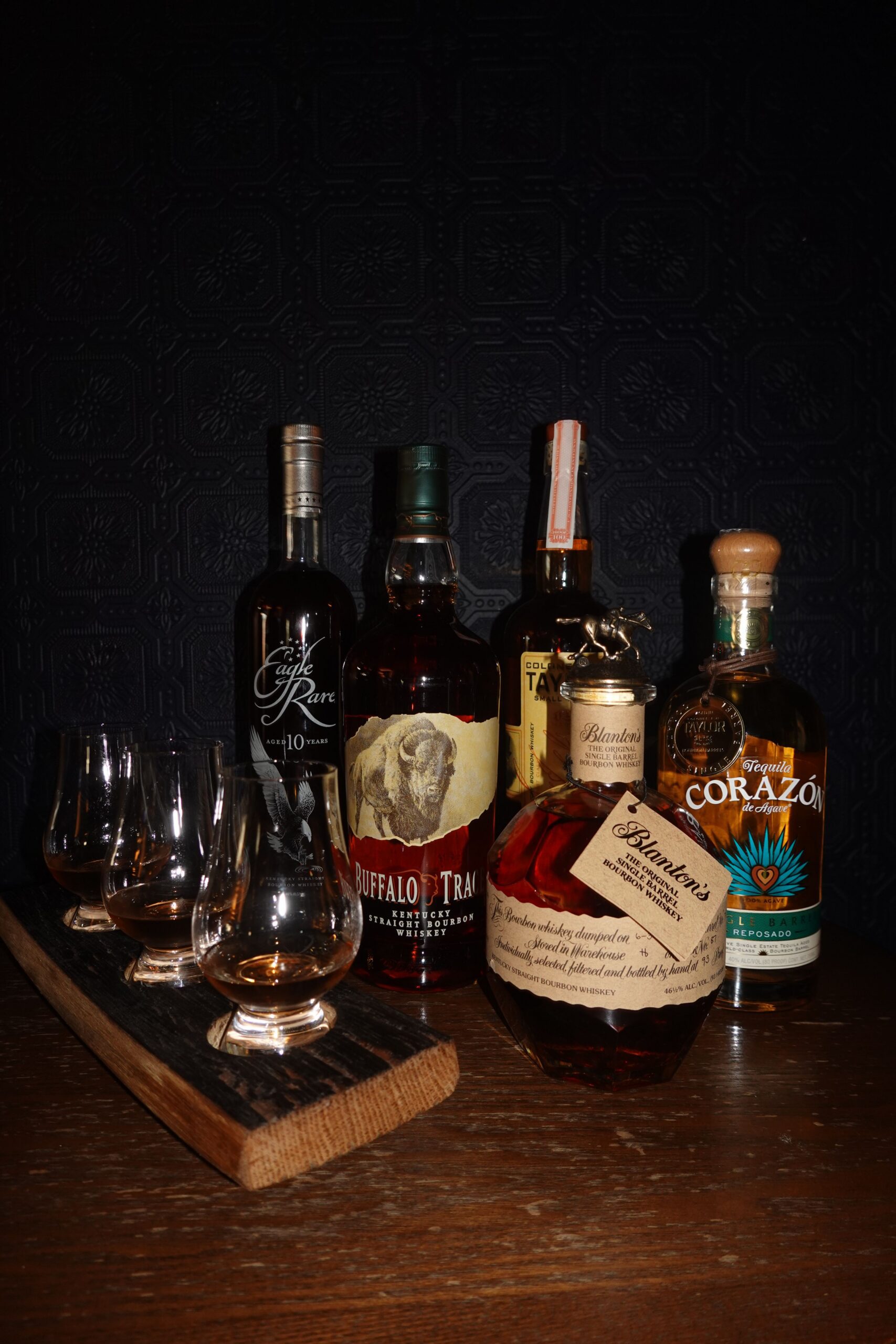Five bottles of whiskey and tequila are arranged on a wooden surface, with four filled tasting glasses on a wooden tray in front—perfect for memorable events—set against a dark textured background.