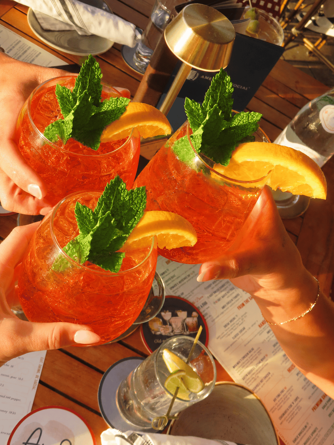 Three hands hold orange cocktails garnished with mint and orange slices above a wooden table set with menus, a glass of water with lime, and tableware, in warm, golden sunlight.