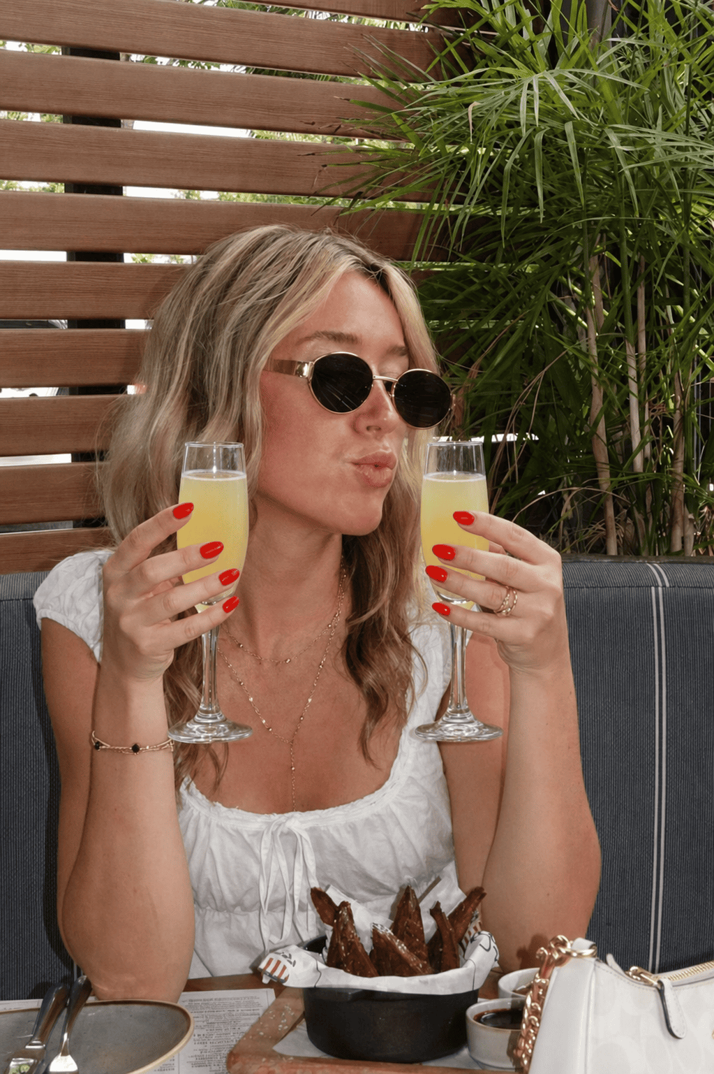 A woman with blonde hair and sunglasses holds two mimosas, making a kissing face. She sits at an outdoor table with food, including chicken wings, and wears a white top with gold jewelry.