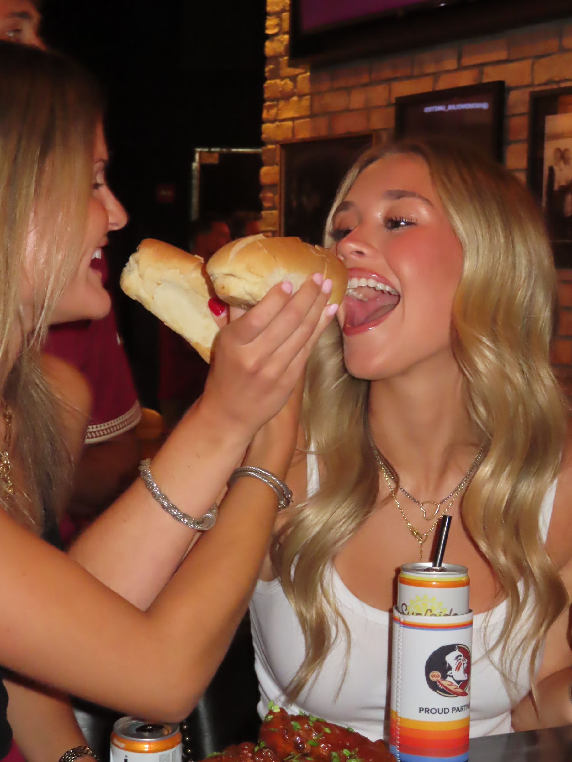 Two women smiling at a table; one holds two sandwich halves near the other’s mouth as she laughs. A drink can and food are visible on the table in front of them.
