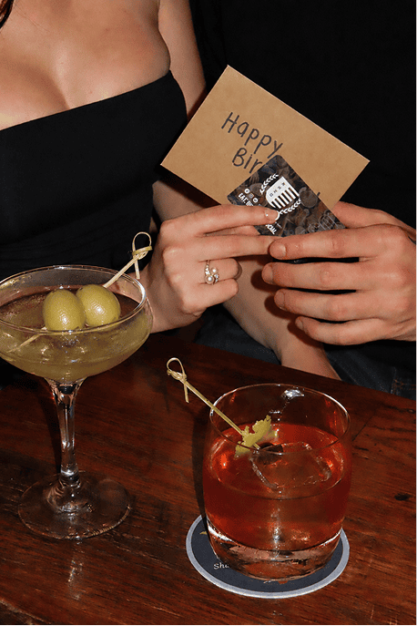 Two people sit at a table with cocktails. One person holds a “Happy Birthday” card and a gift card. The other wears a black strapless top. Two drinks with garnishes are on the wooden table.